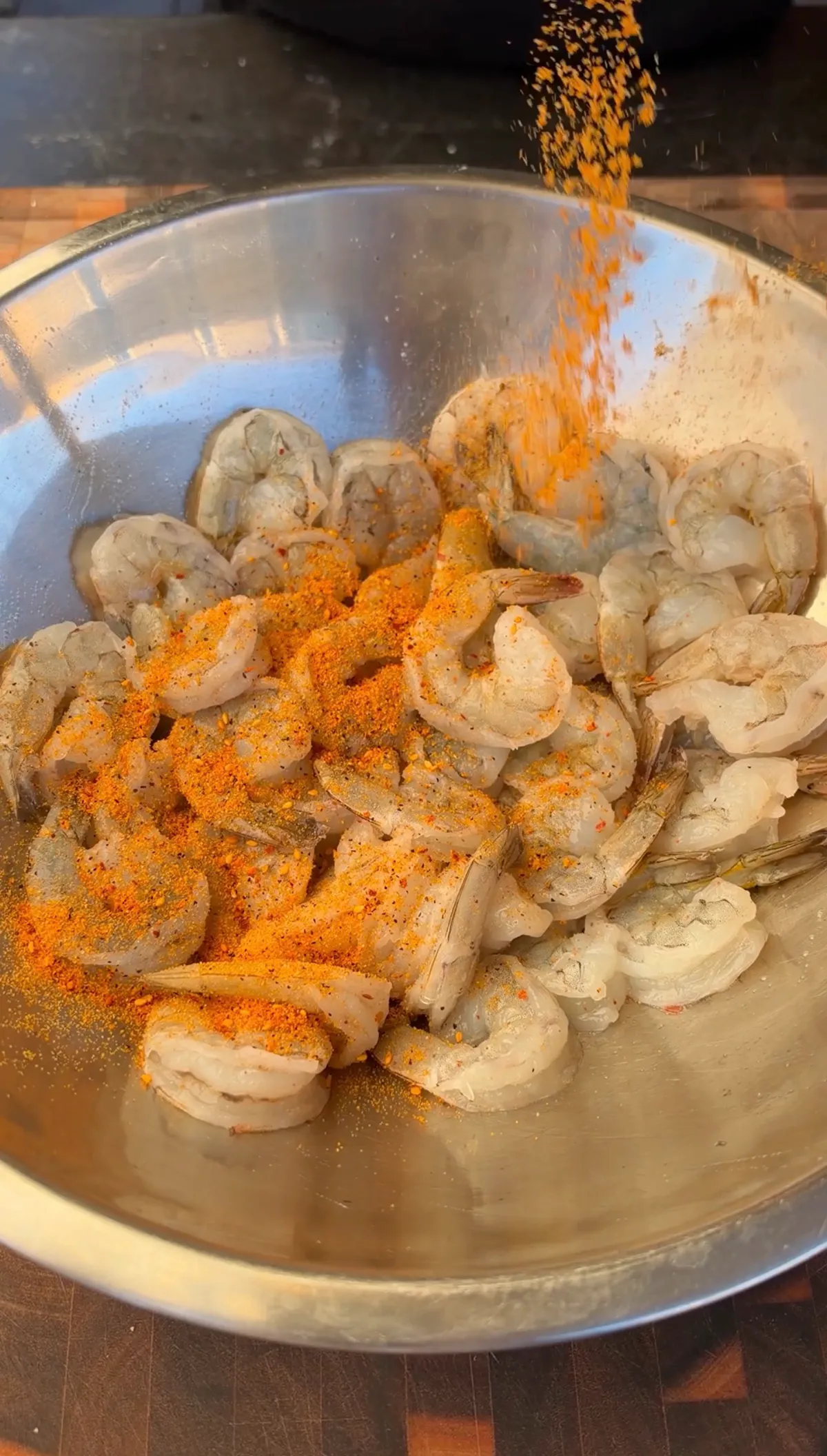 Seafood seasoning being poured over peeled raw shrimp in a stainless steel mixing bowl