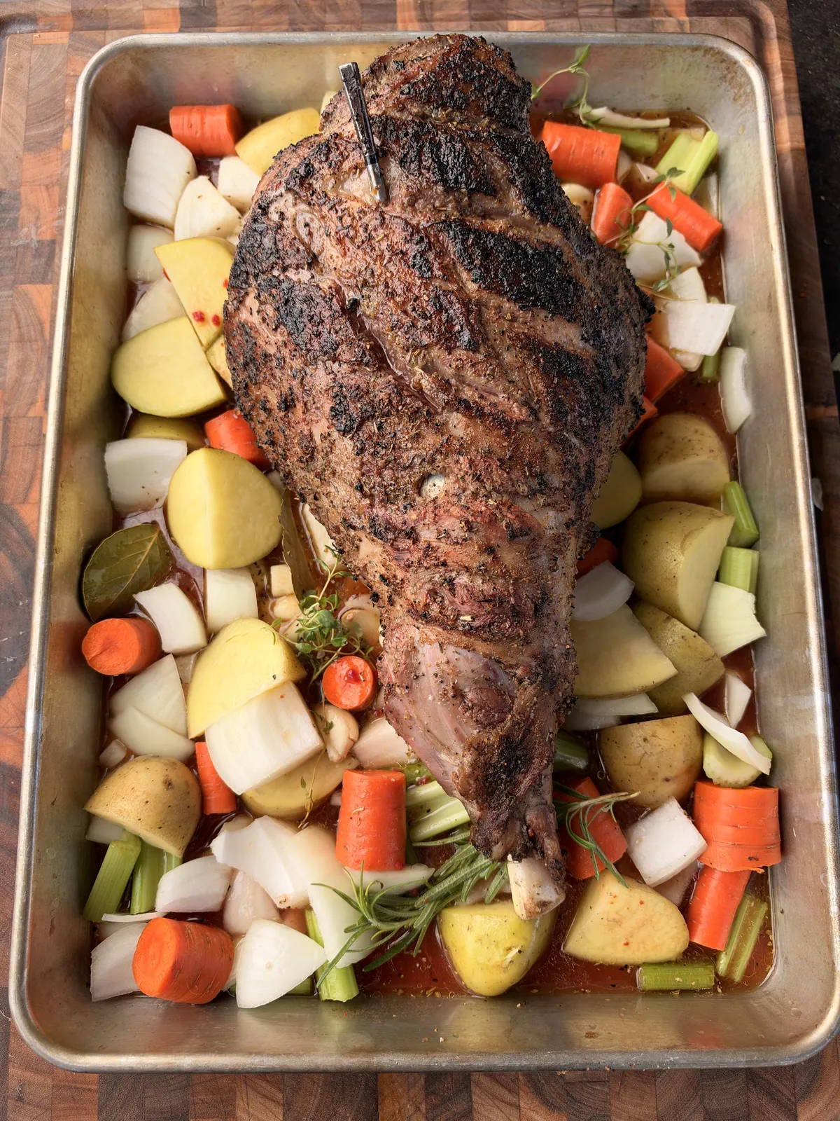 Seared Greek-style leg of lamb with MEATER thermometer resting on Yukon Gold potatoes, carrots, celery, and onions in a roasting pan