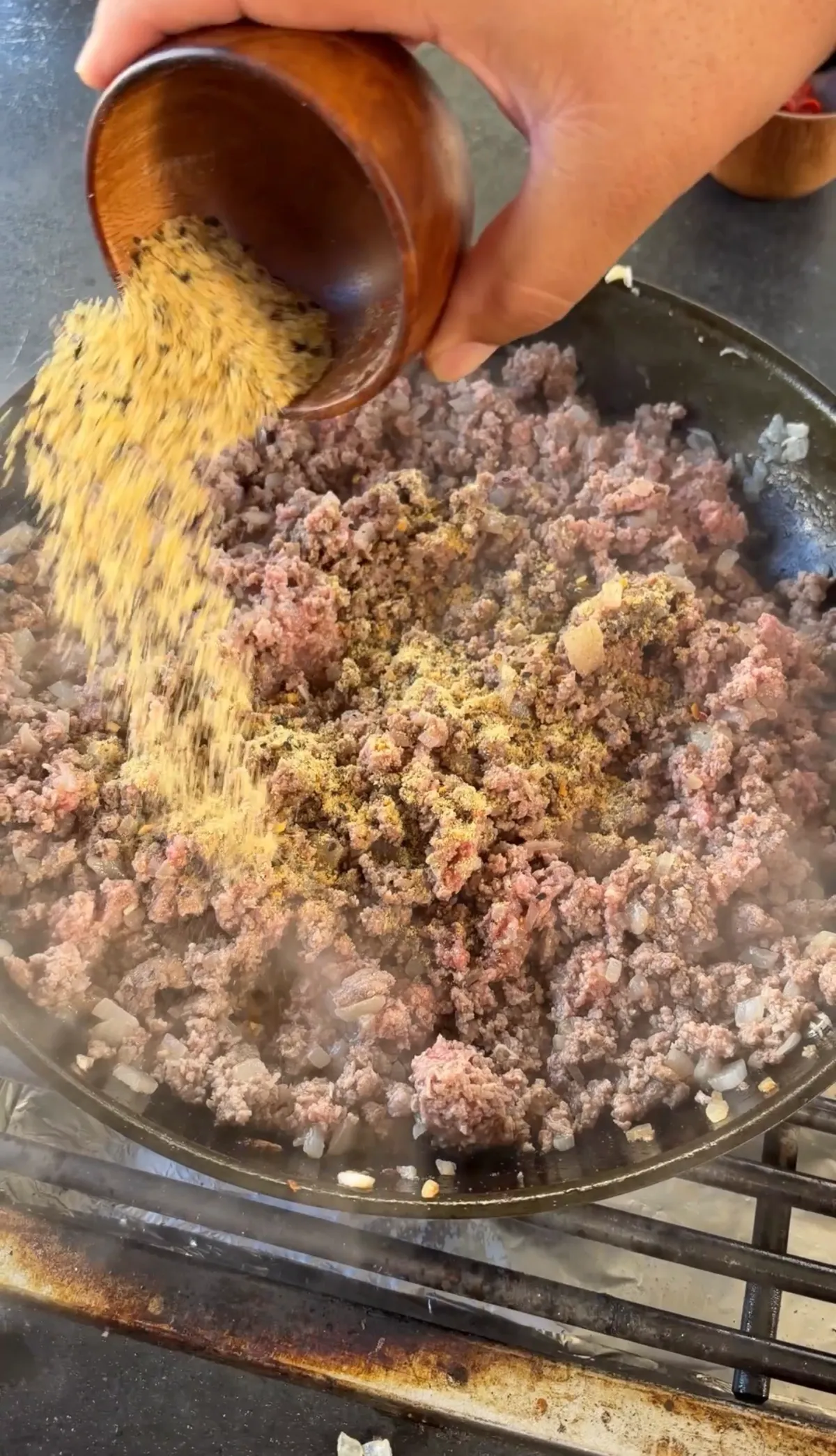 Burger seasonings being poured from wooden bowl onto 85/15 ground beef and onions cooking in cast iron skillet