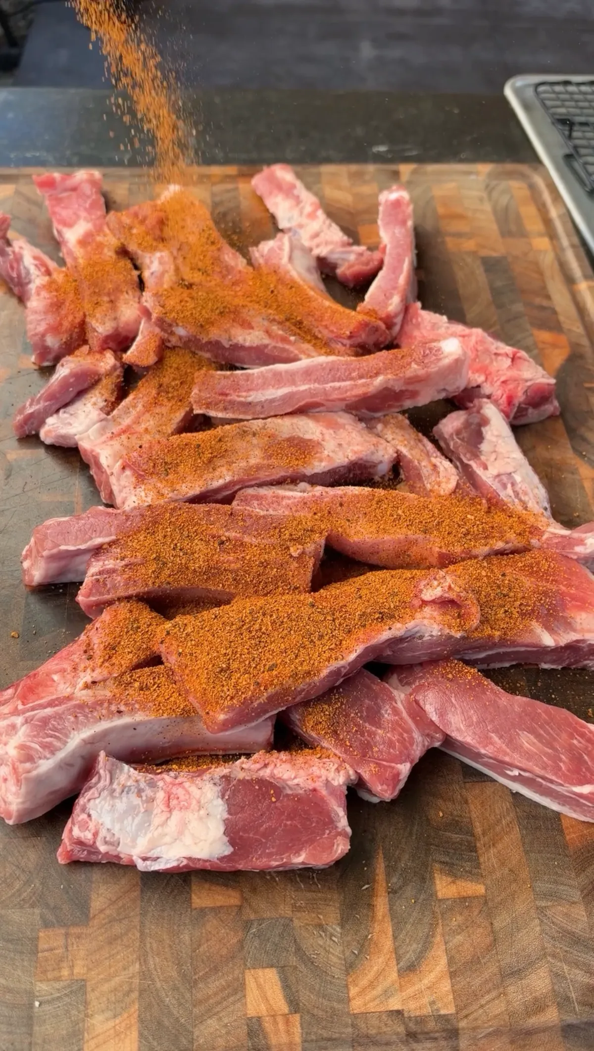 Raw individual St. Louis-style pork ribs on a cutting board being generously coated with cherry barbecue seasoning rub