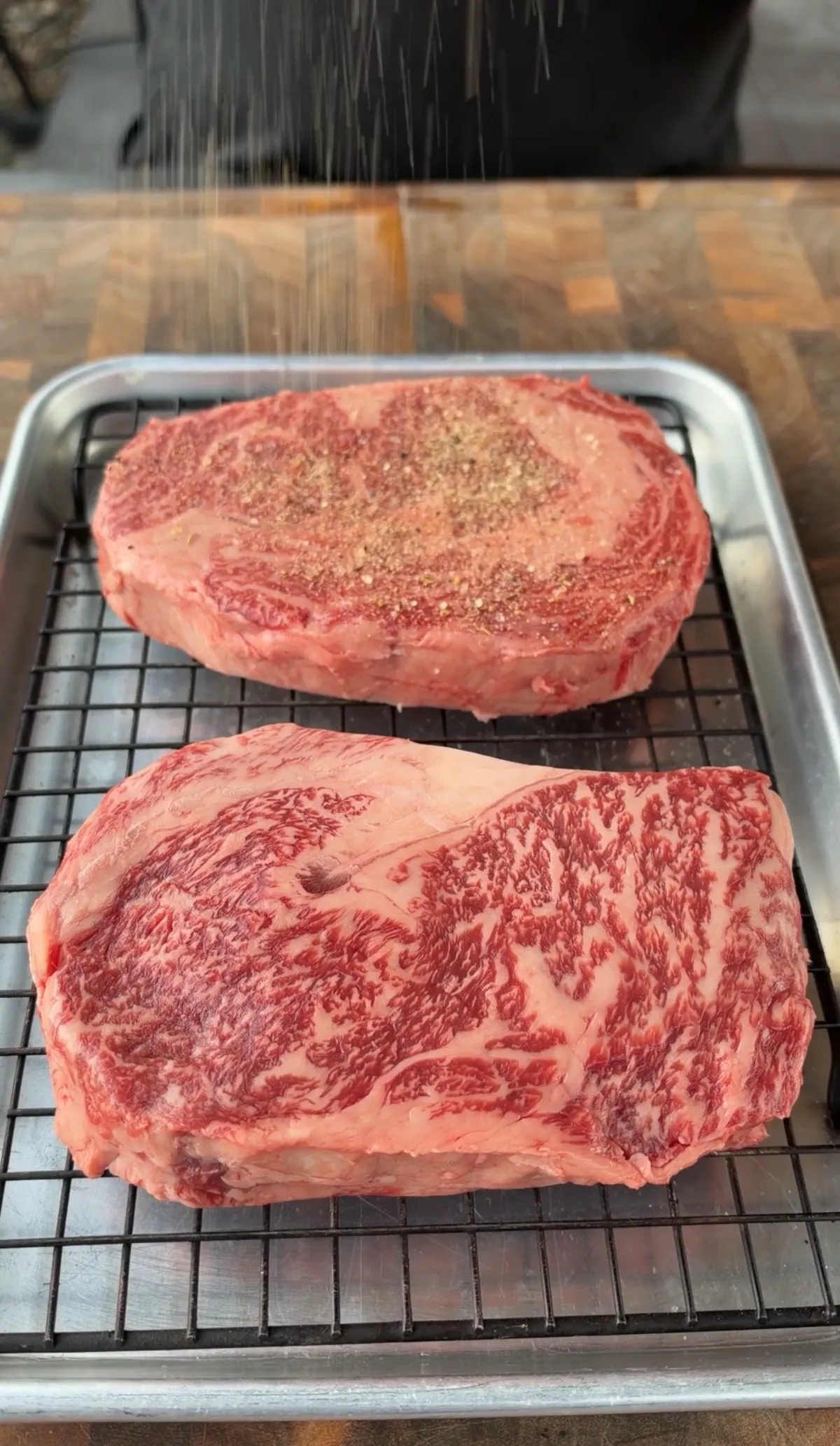 Two raw ribeye steaks on a wire rack being seasoned with steak seasoning before searing