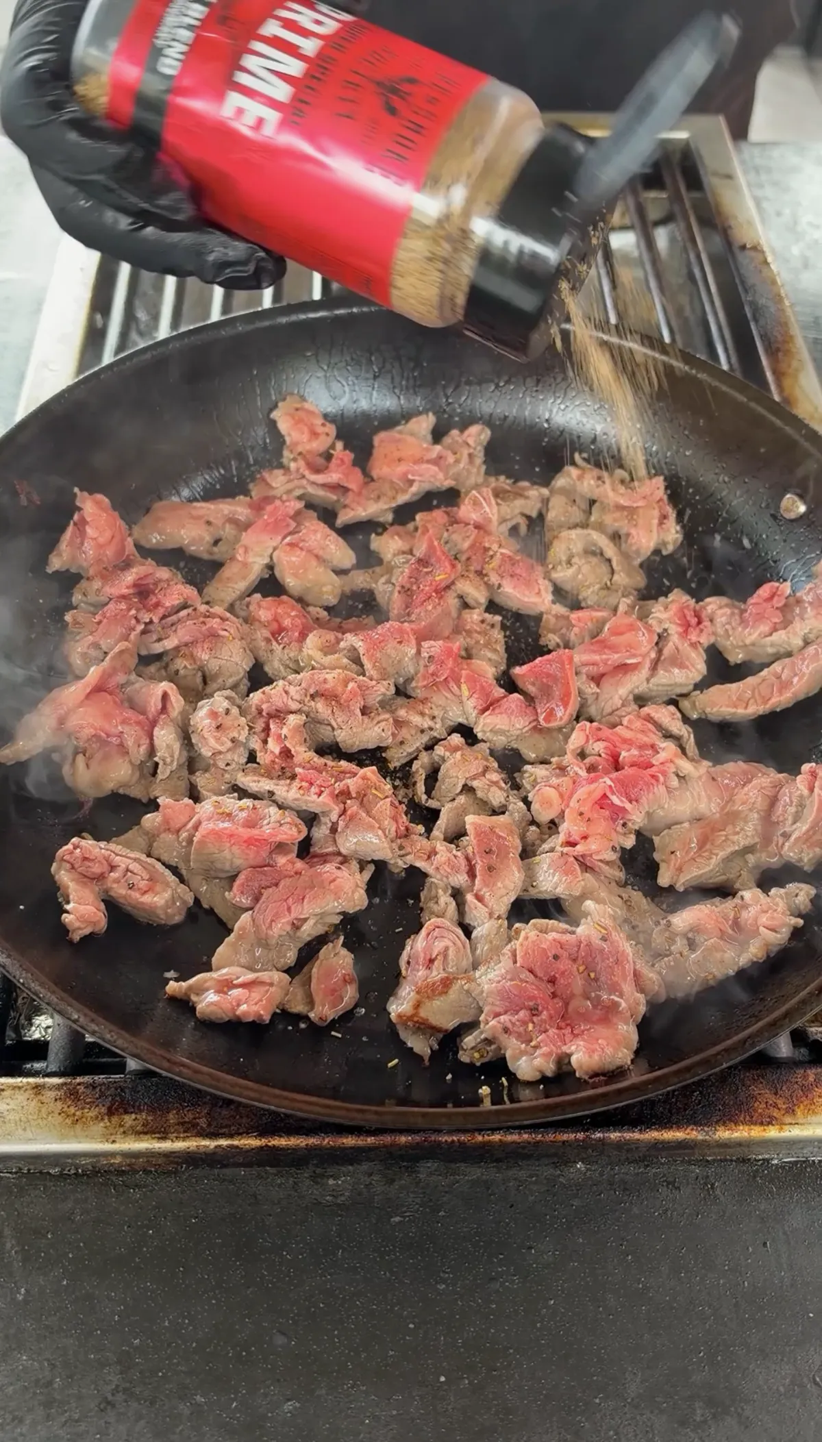 Thinly sliced ribeye steak strips cooking in a black skillet being seasoned with steak seasoning from a red shaker