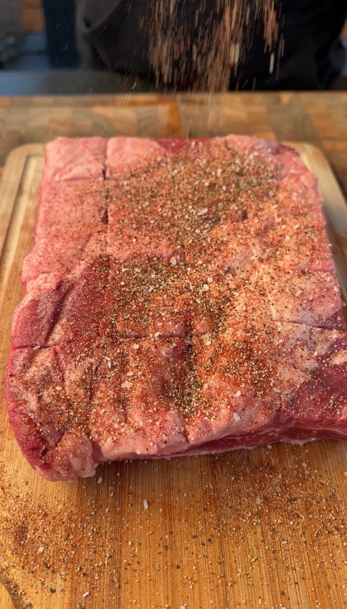 Raw beef short ribs being seasoned with spice rub on a wooden cutting board, with seasoning falling from above