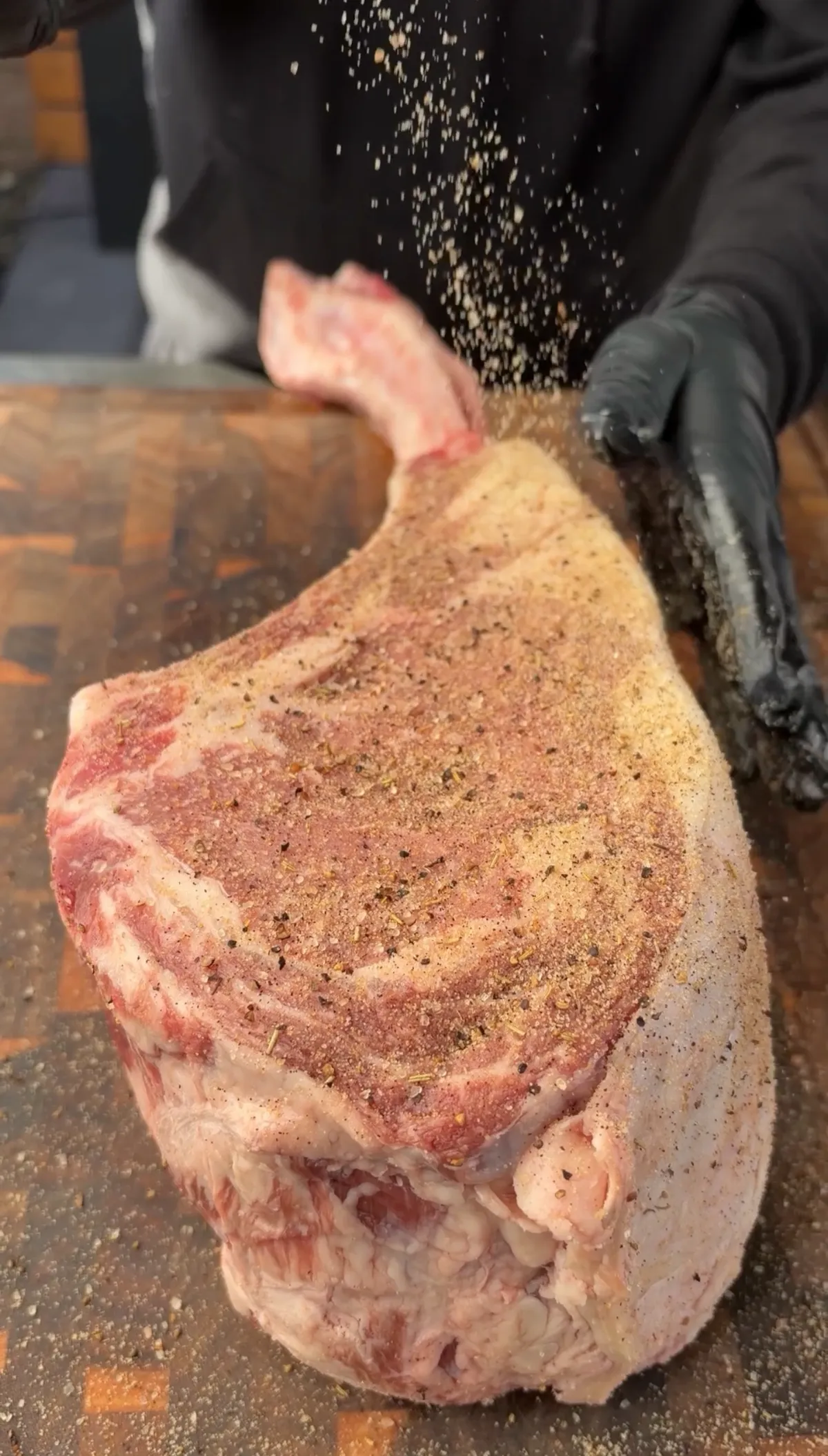 Steak seasoning being sprinkled over a raw two-bone prime rib tomahawk on a wooden cutting board