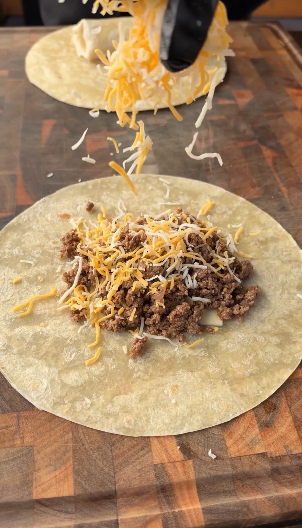 Shredded Colby Jack cheese being poured over seasoned 85/15 ground beef on a flour tortilla for a fried burrito