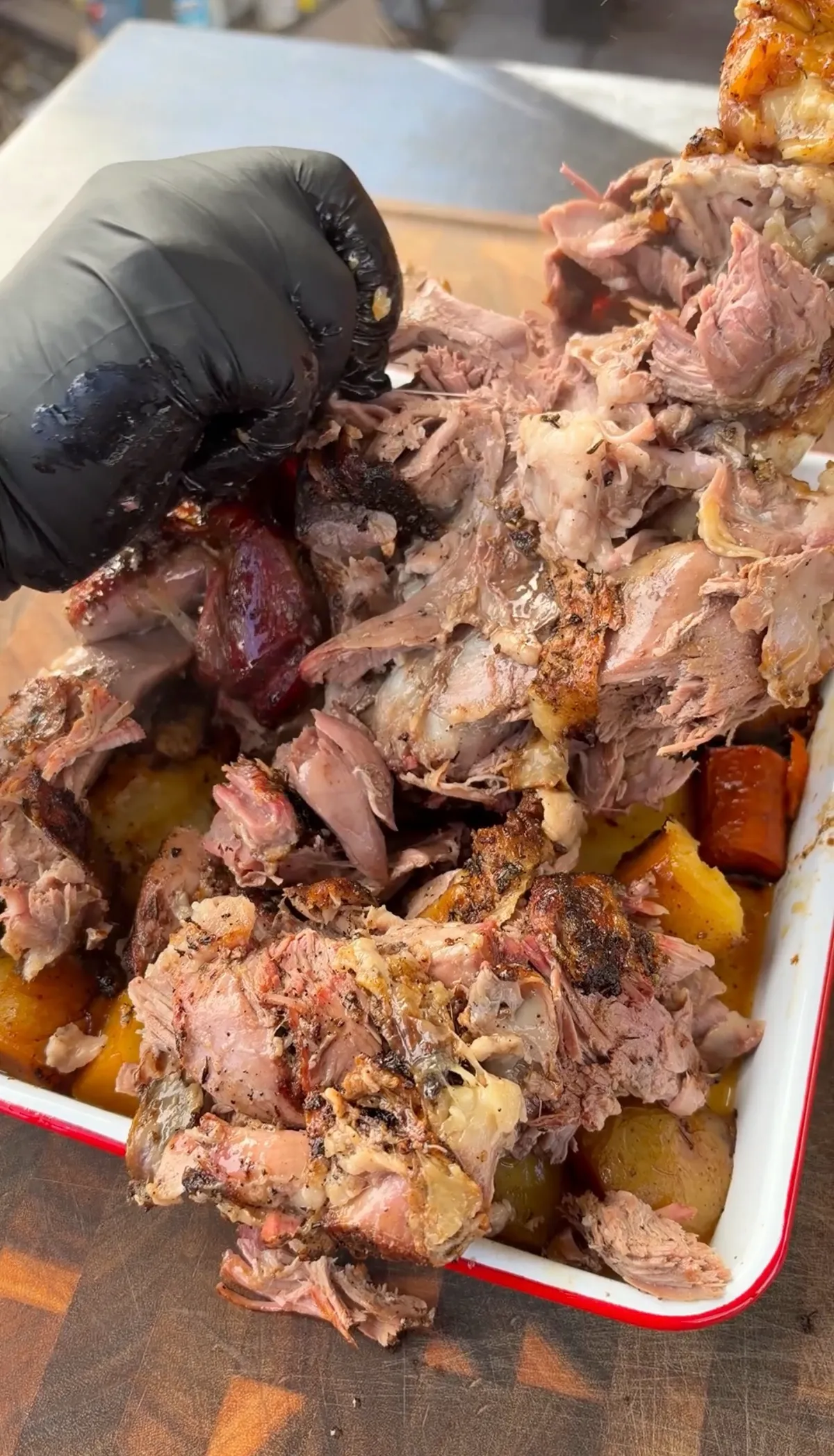 Black-gloved hands shredding fall-apart tender braised leg of lamb over Yukon Gold potatoes and carrots in white roasting tray