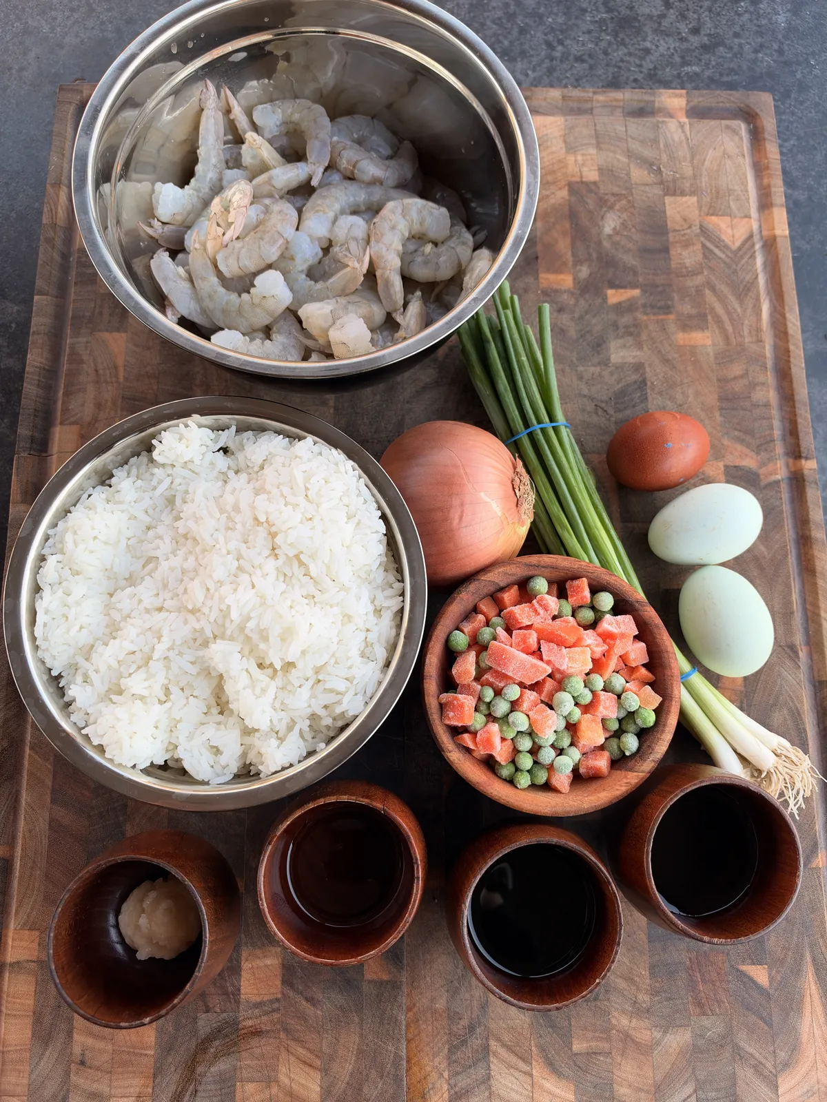 Shrimp fried rice ingredients prepped on a wooden board: raw shrimp, day-old rice, eggs, yellow onion, frozen peas and carrots, green onions, and sauce mix