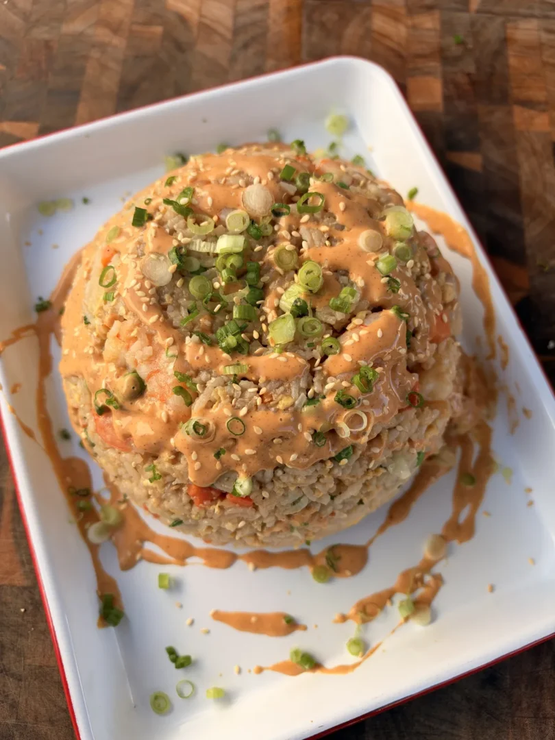 Domed shrimp fried rice topped with yum yum sauce, sesame seeds, and sliced green onions on a white plate