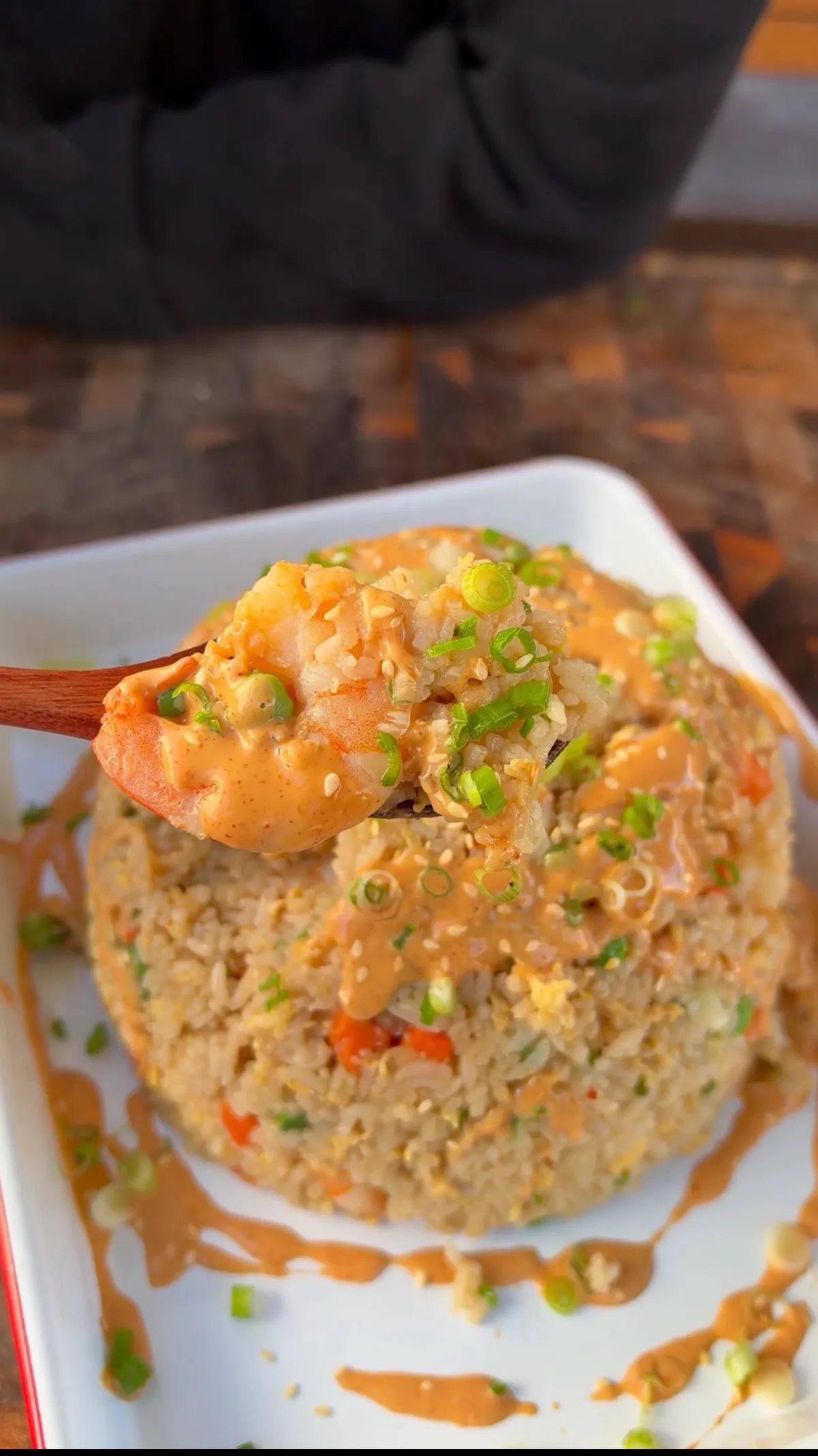 Wooden spoon lifting shrimp fried rice with yum yum sauce drizzle and sliced green onions on white plate