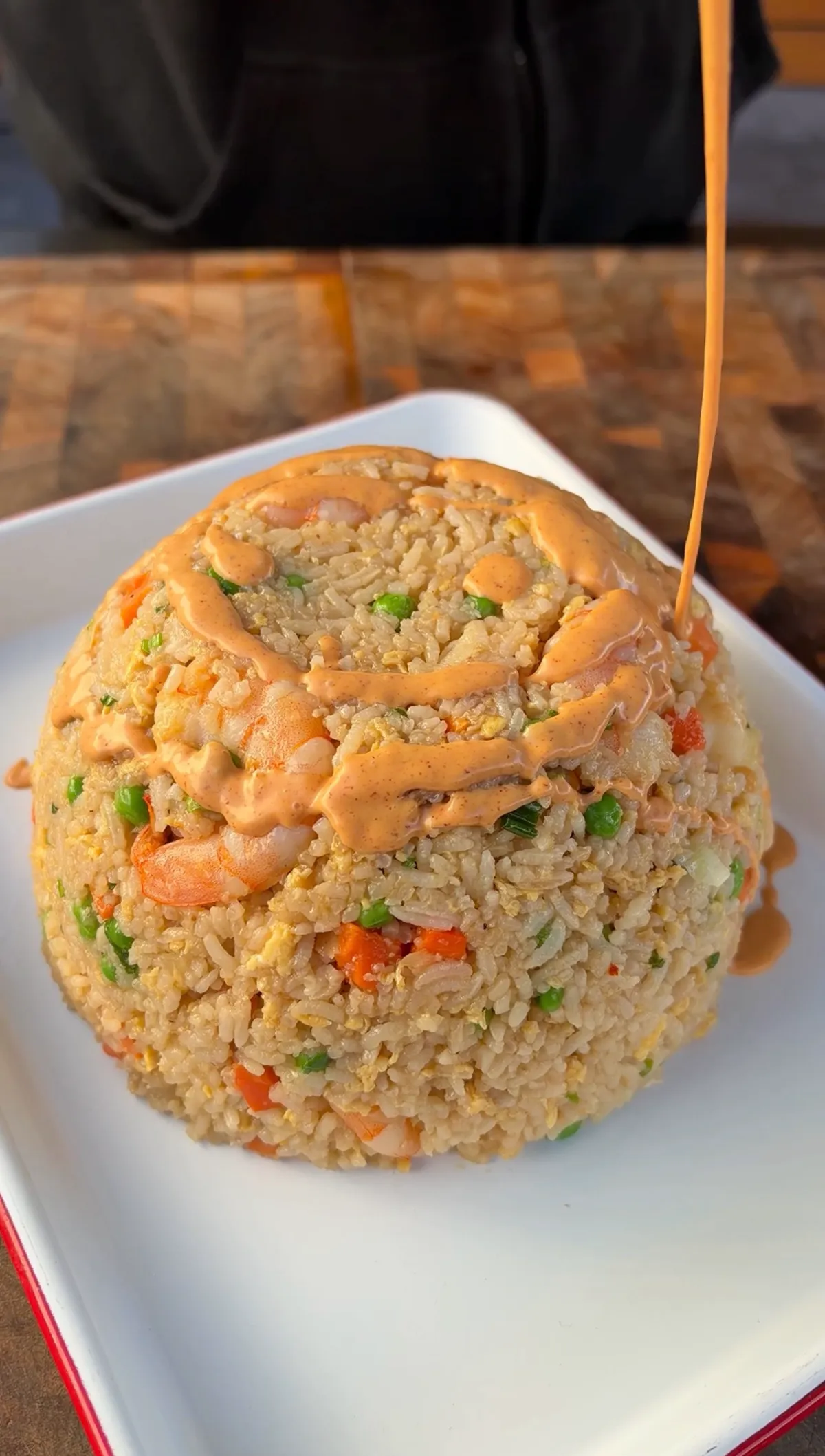 Domed shrimp fried rice with peas, carrots, and egg on a white plate, topped with creamy yum yum sauce being poured