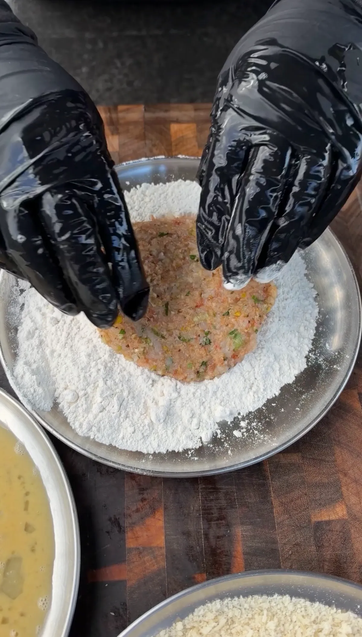 Gloved hands coating a raw shrimp burger patty in all-purpose flour in a metal bowl at the breading station