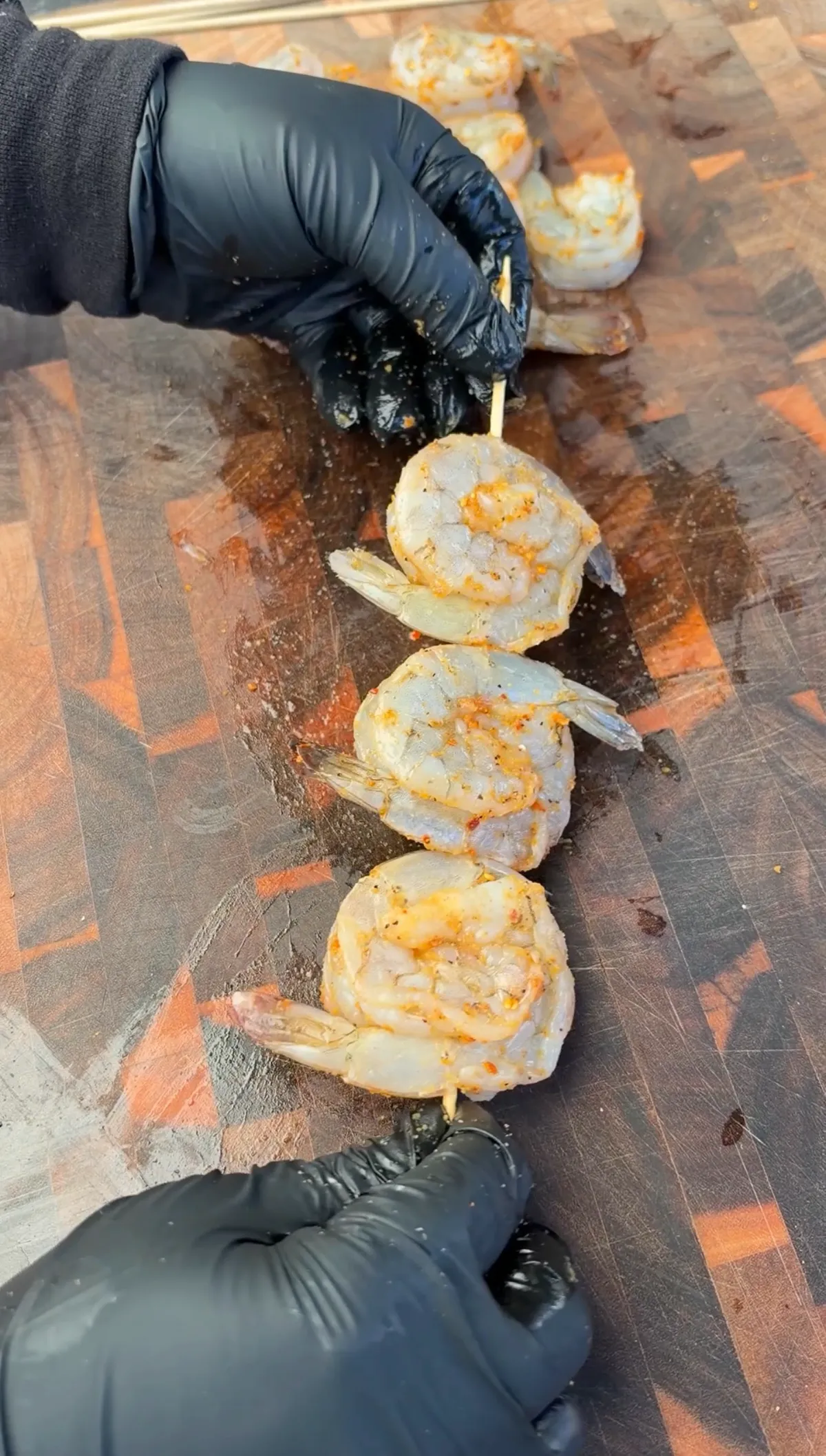 Gloved hands threading seasoned shrimp in overlapping yin-yang shape onto wooden skewers on a cutting board