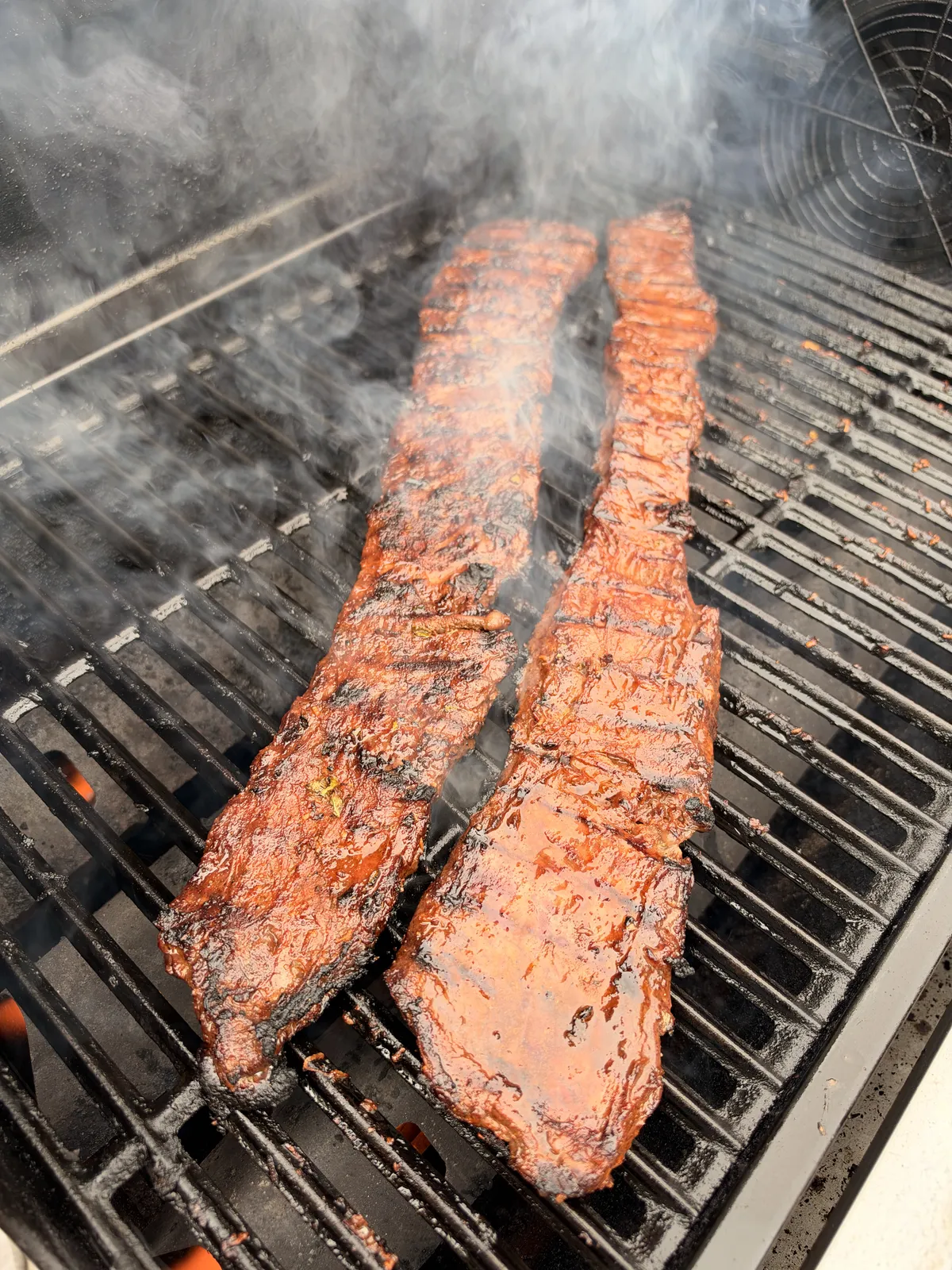 Two strips of marinated skirt steak grilling over high heat with smoke rising, developing a deep char on a BBQ grill