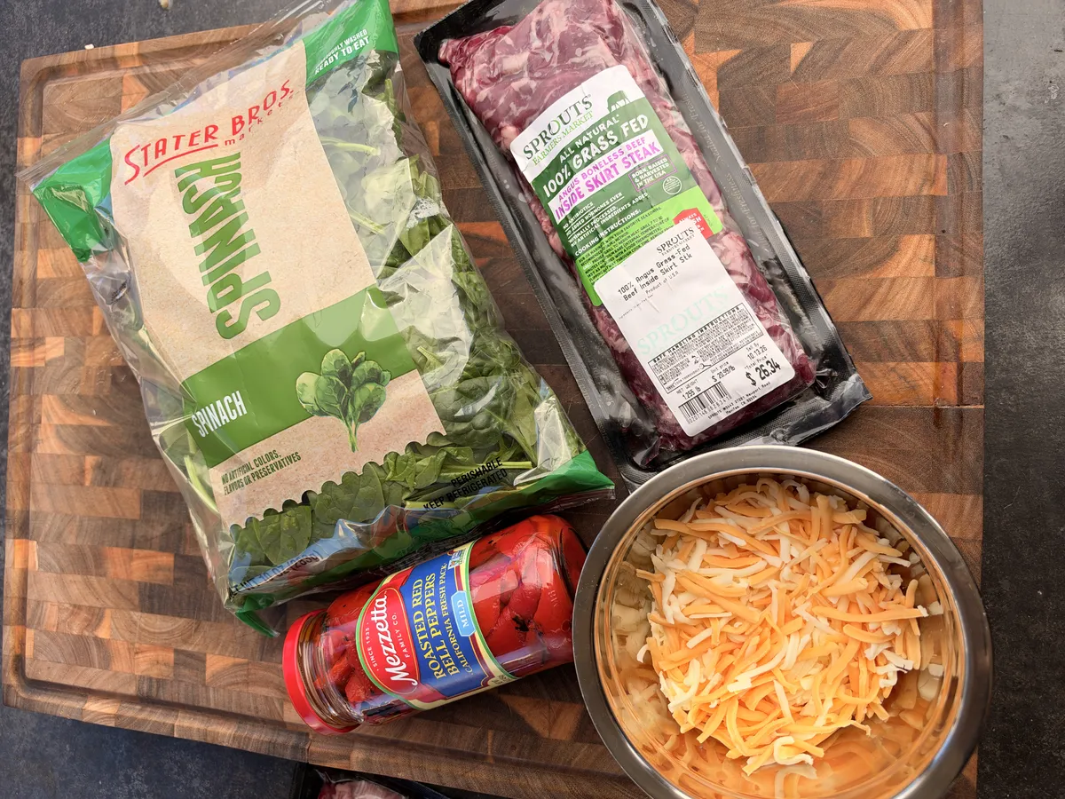 Skirt steak pinwheel ingredients: grass-fed skirt steak, fresh spinach, roasted red bell peppers, and shredded Colby Jack cheese on a wood cutting board