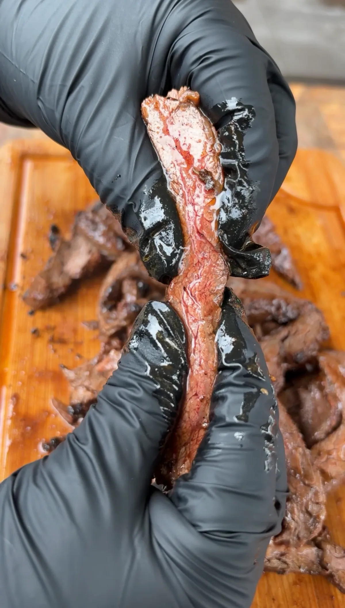 Gloved hands pulling apart a slice of grilled skirt steak to show juicy interior and tender grain on a wooden cutting board