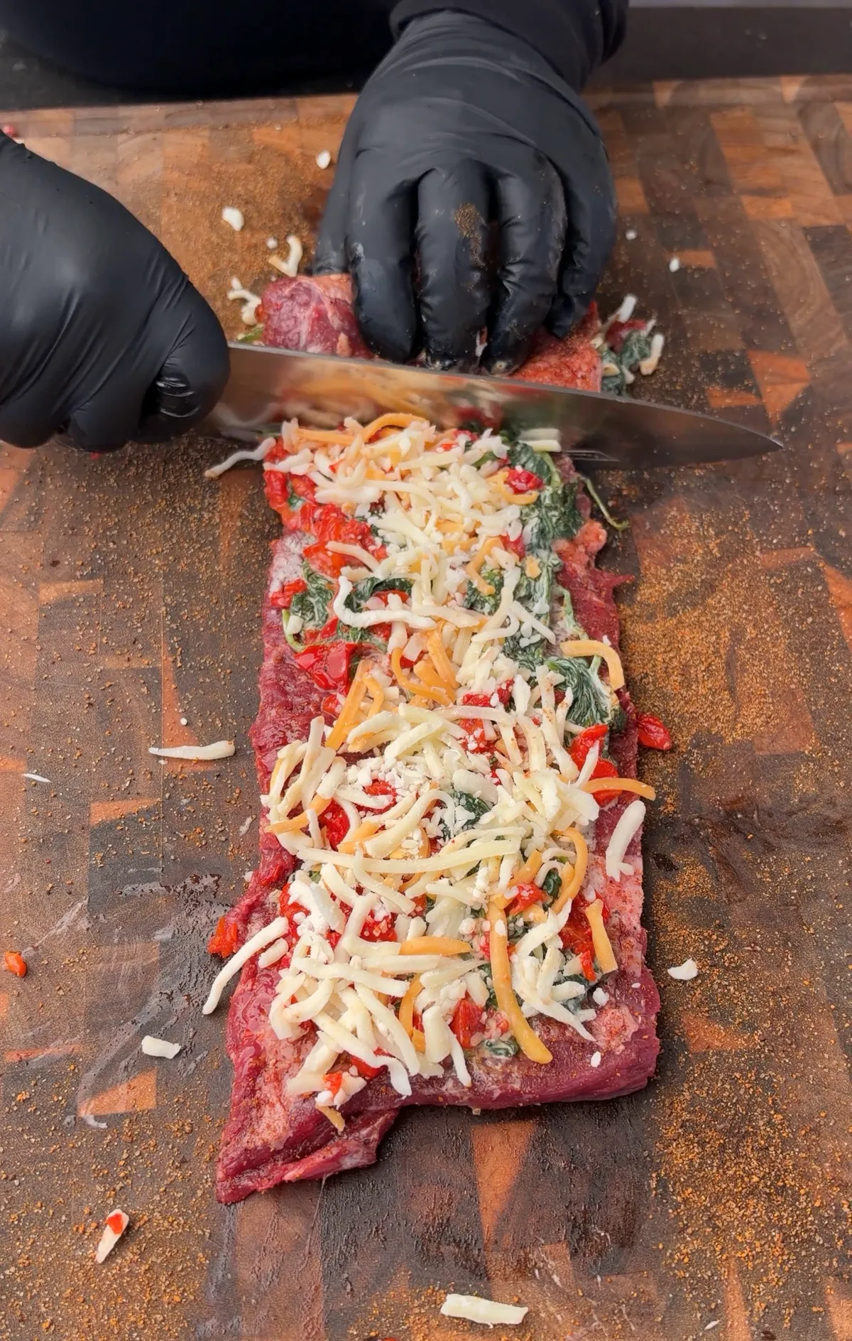 Hands in black gloves rolling skirt steak filled with creamy spinach, roasted red bell peppers, and grated Colby Jack cheese on a cutting board