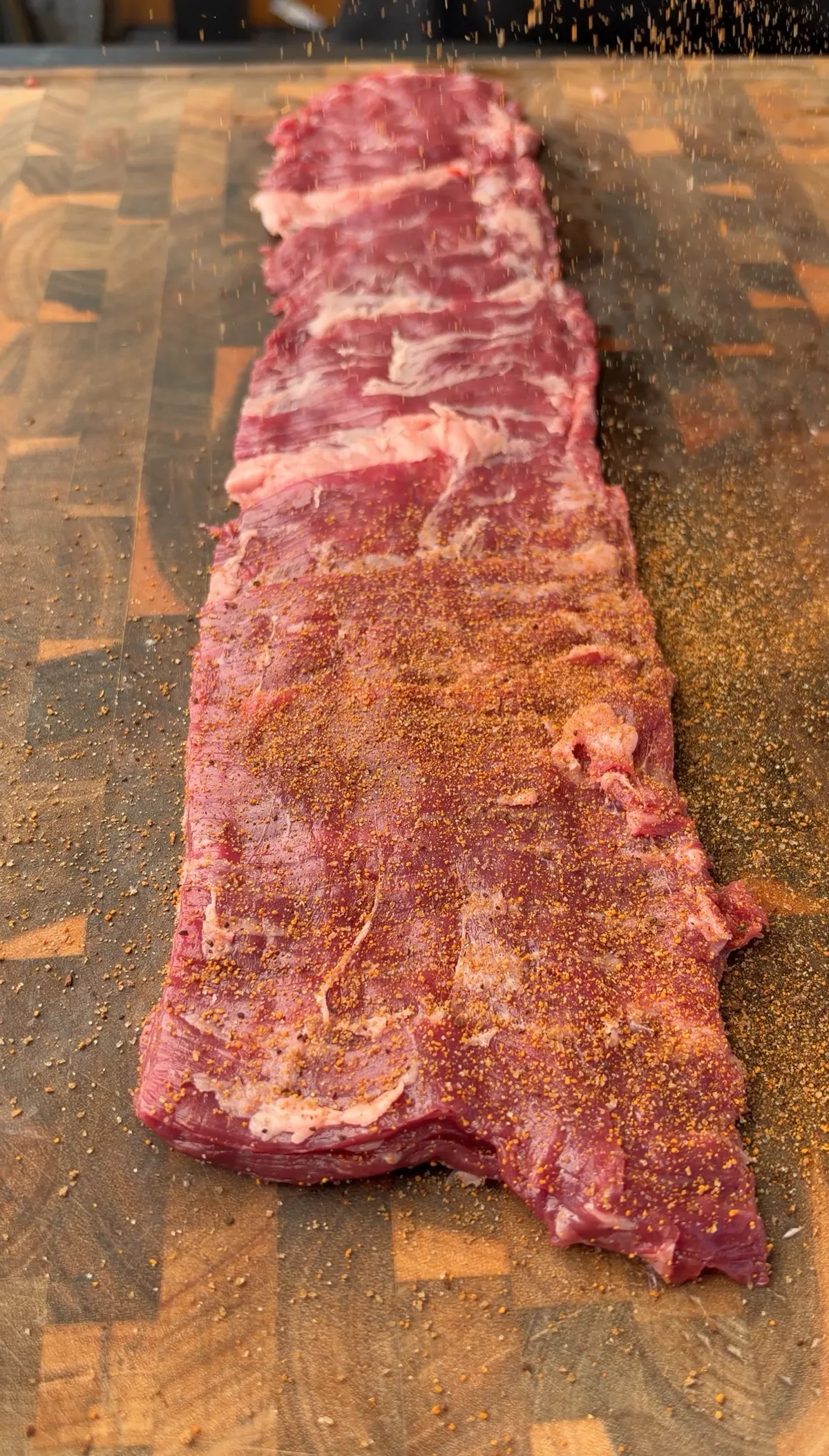 Raw skirt steak being generously seasoned with steak seasoning on a wooden cutting board