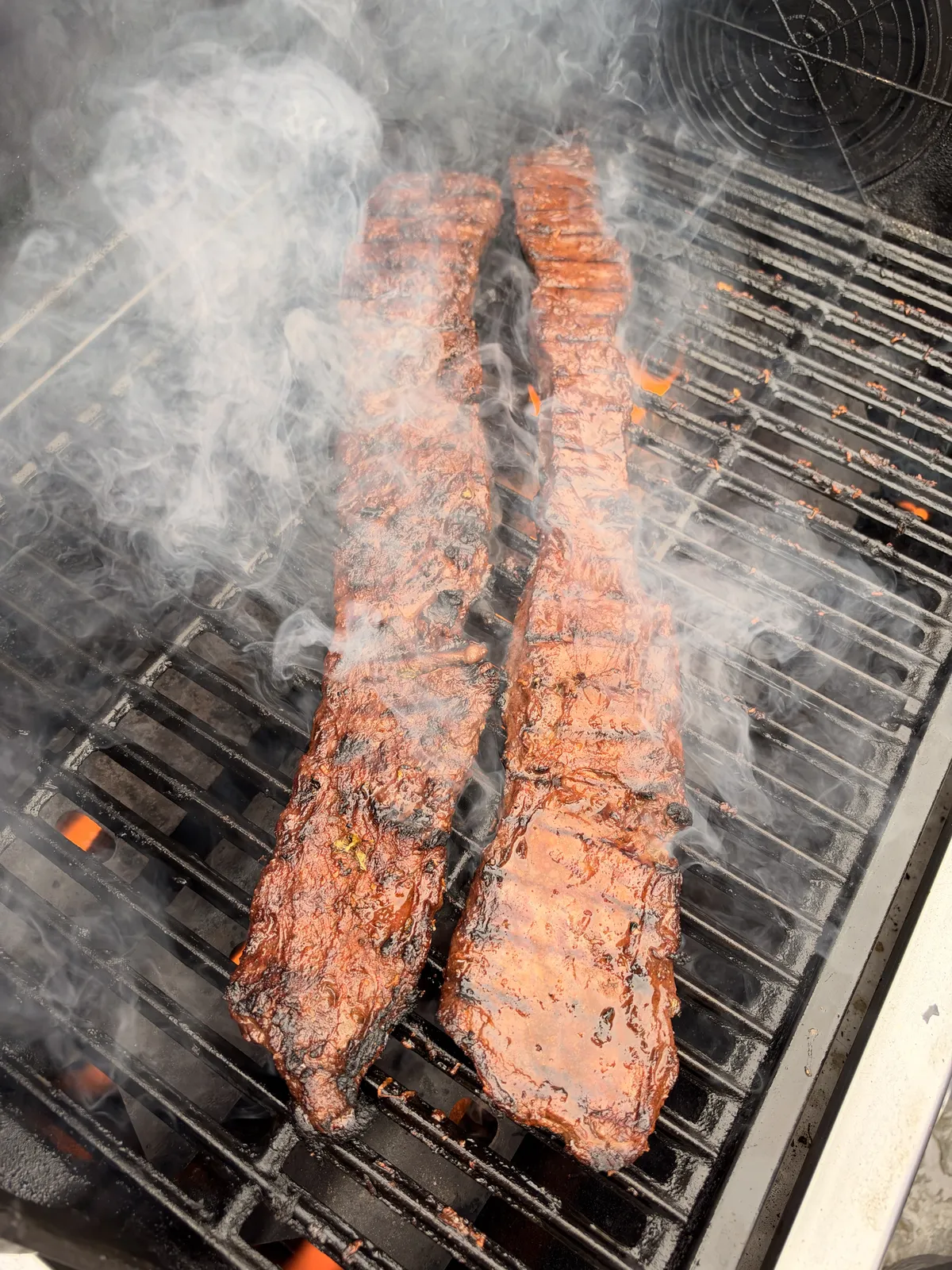Two marinated skirt steaks grilling over high heat with smoke rising from charred, saucy grill grates