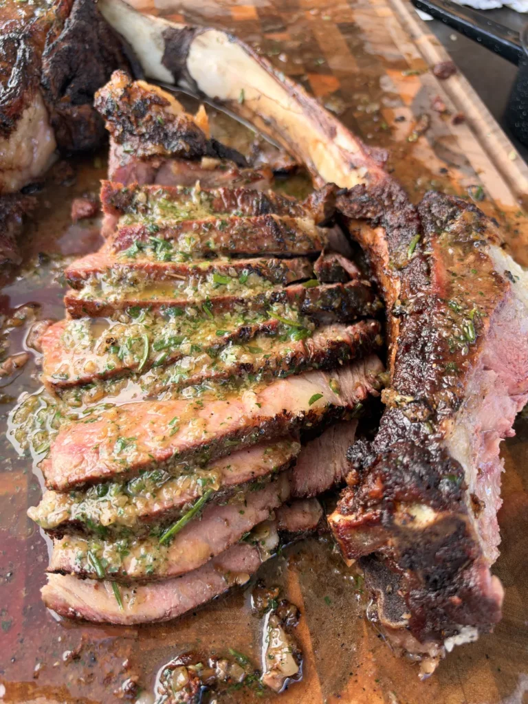 Sliced two-bone prime rib tomahawk with roasted garlic and onion compound butter melting over each slice on a cutting board