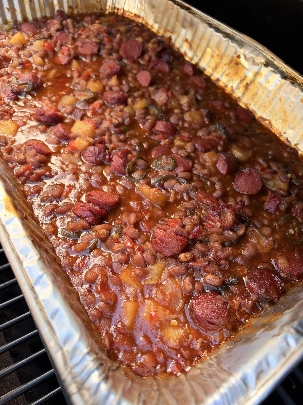 Smoked BBQ baked beans with sliced hot dogs, jalapeños, pineapple, and bacon in a foil pan on smoker grates