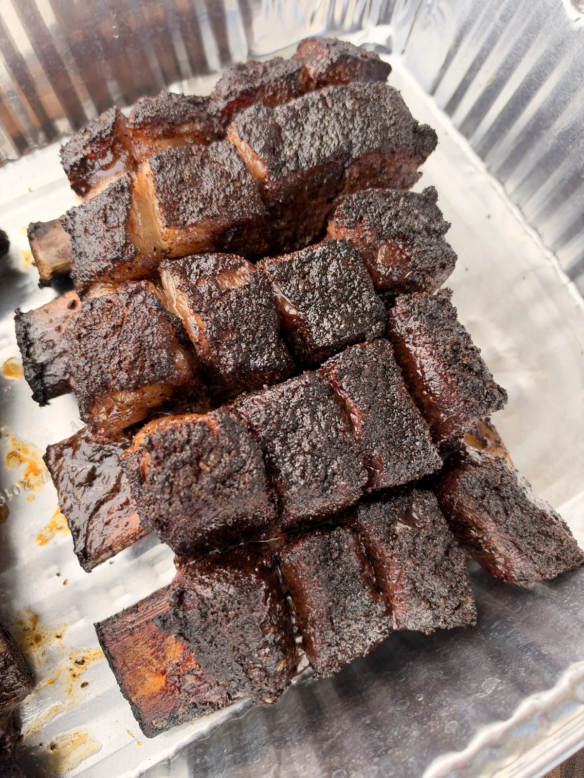 Heavily smoked beef short rib cubes with dark bark crust piled high in a disposable aluminum foil pan