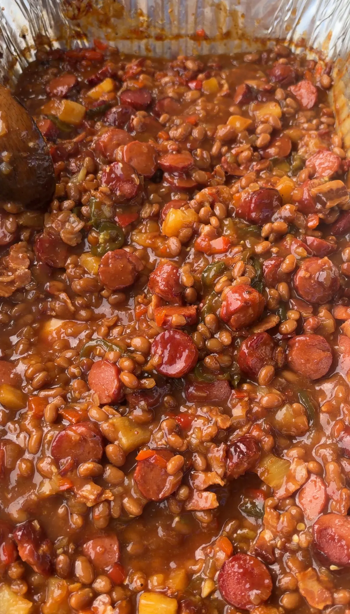 Smoked BBQ baked beans with sliced hot dogs, jalapeños, and bacon in a glass baking dish being stirred