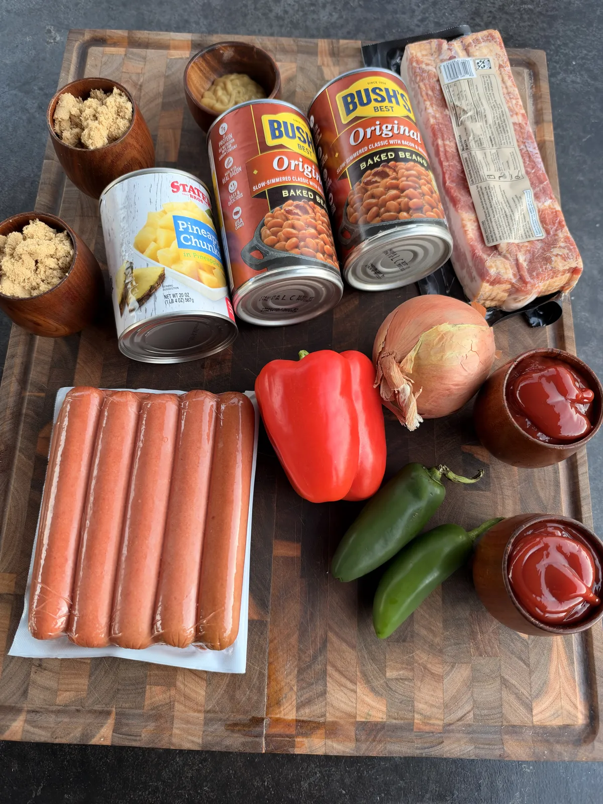 Smoked BBQ baked beans ingredients: Bush's baked beans, bacon, hot dogs, red bell pepper, jalapeños, onion, pineapple chunks, brown sugar, and ketchup on a wooden cutting board