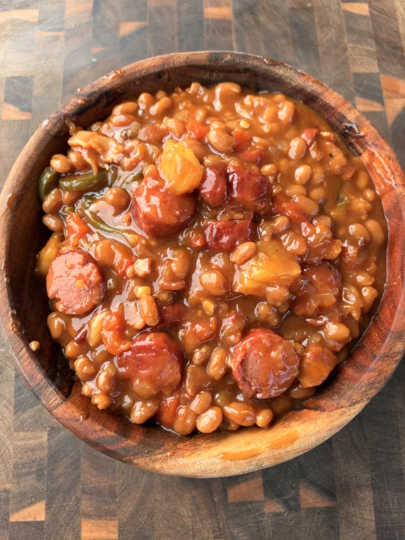 Smoked BBQ baked beans with sliced hot dogs, jalapeños, and pineapple in a rustic wooden bowl