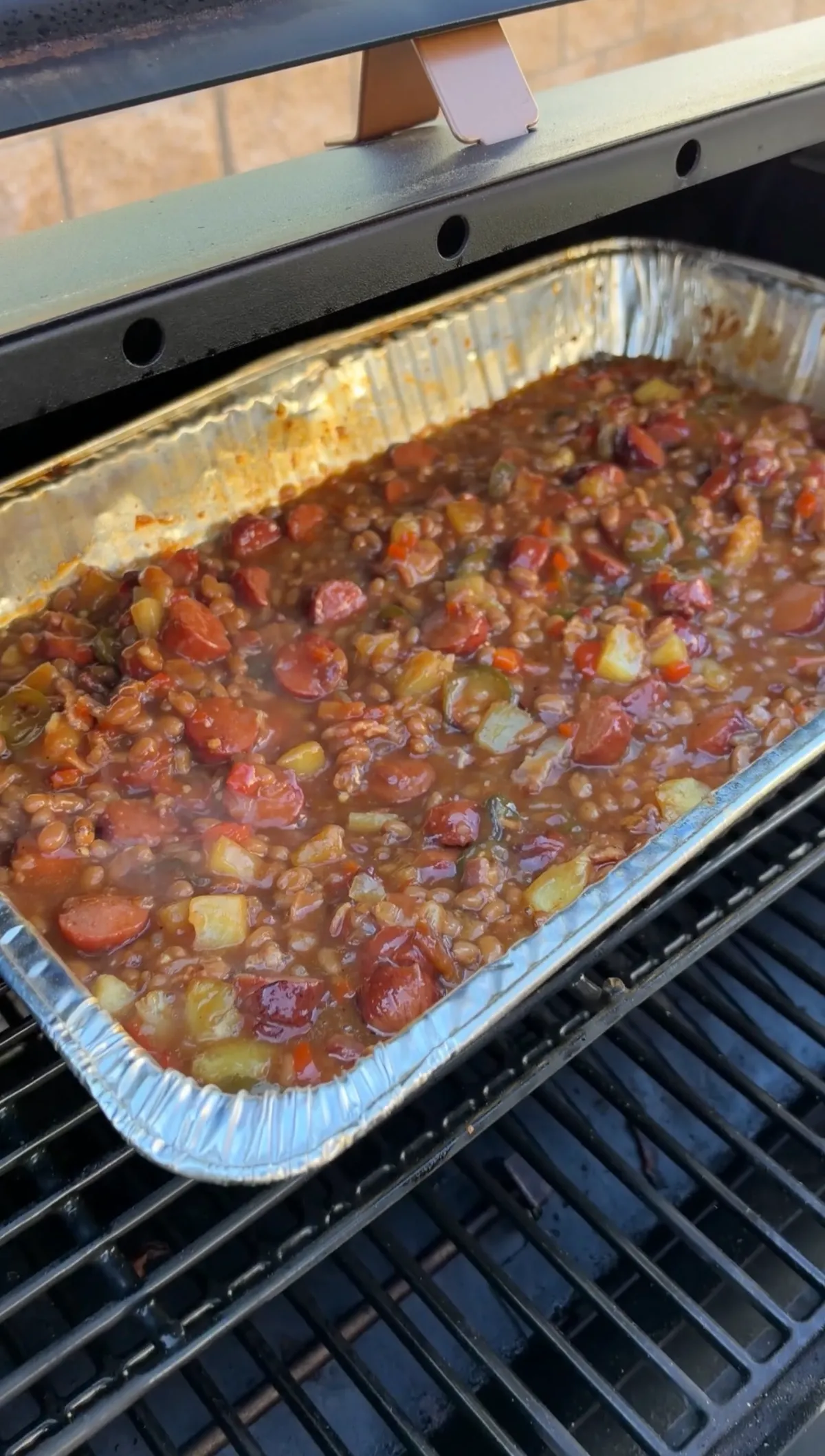 Smoked BBQ baked beans with hot dog rounds, pineapple, and jalapeños bubbling in a foil pan on smoker grates