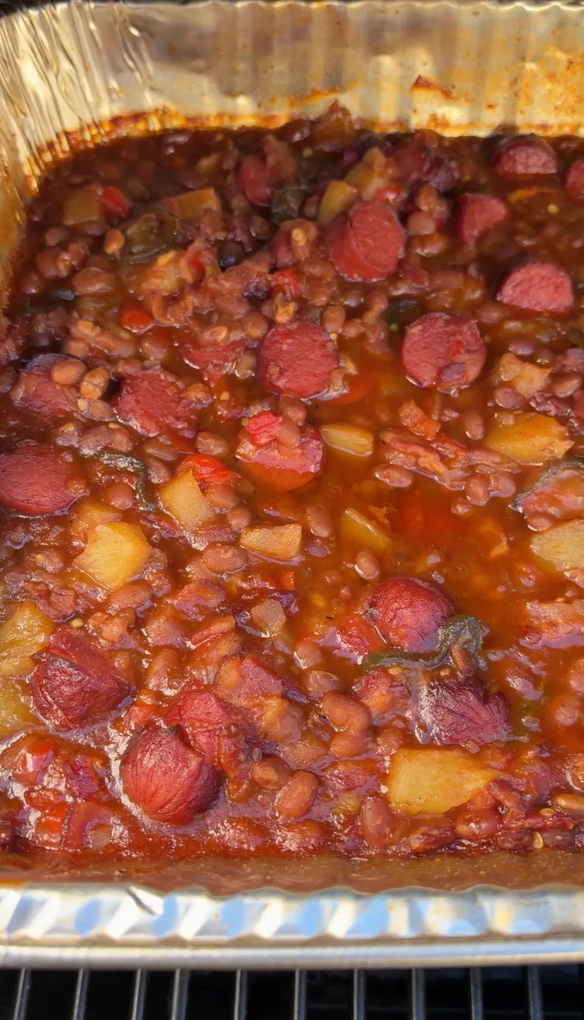 Smoked BBQ baked beans with sliced hot dog rounds, pineapple tidbits, and jalapeños in a foil pan after 3 hours at 250°F