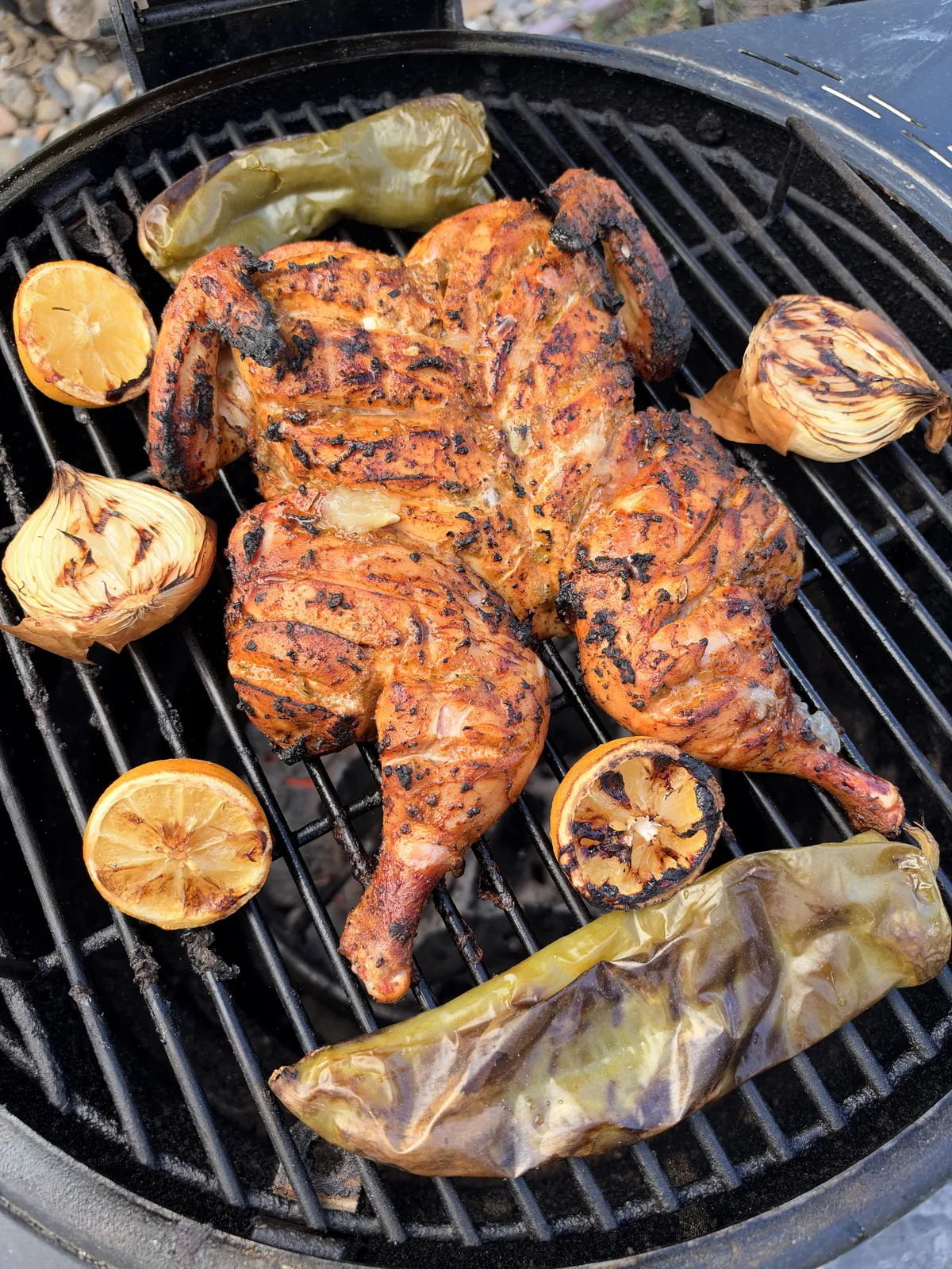Spatchcocked chicken grilled with charred Anaheim peppers, halved lemons, and whole onions on a round charcoal grill