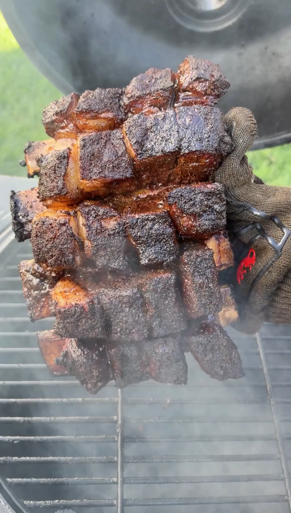 Gloved hand holding a large stack of smoky beef short rib burnt ends with dark bark crust over a smoker grate