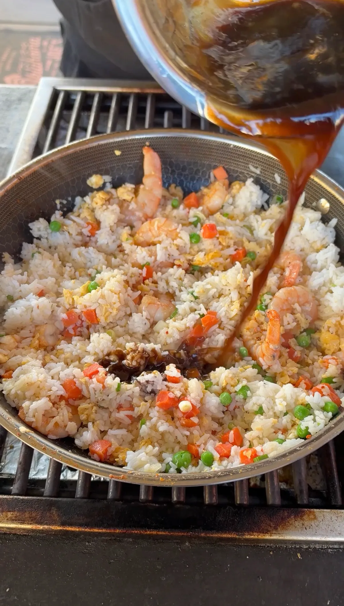 Soy sauce, oyster sauce, and sesame oil mixture being poured over shrimp fried rice with peas and carrots in a wok on a grill