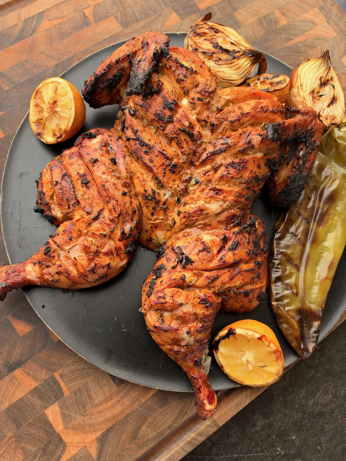 Smoked green chile lime grilled spatchcocked chicken on dark platter with charred lemons, grilled onions, and roasted Anaheim pepper