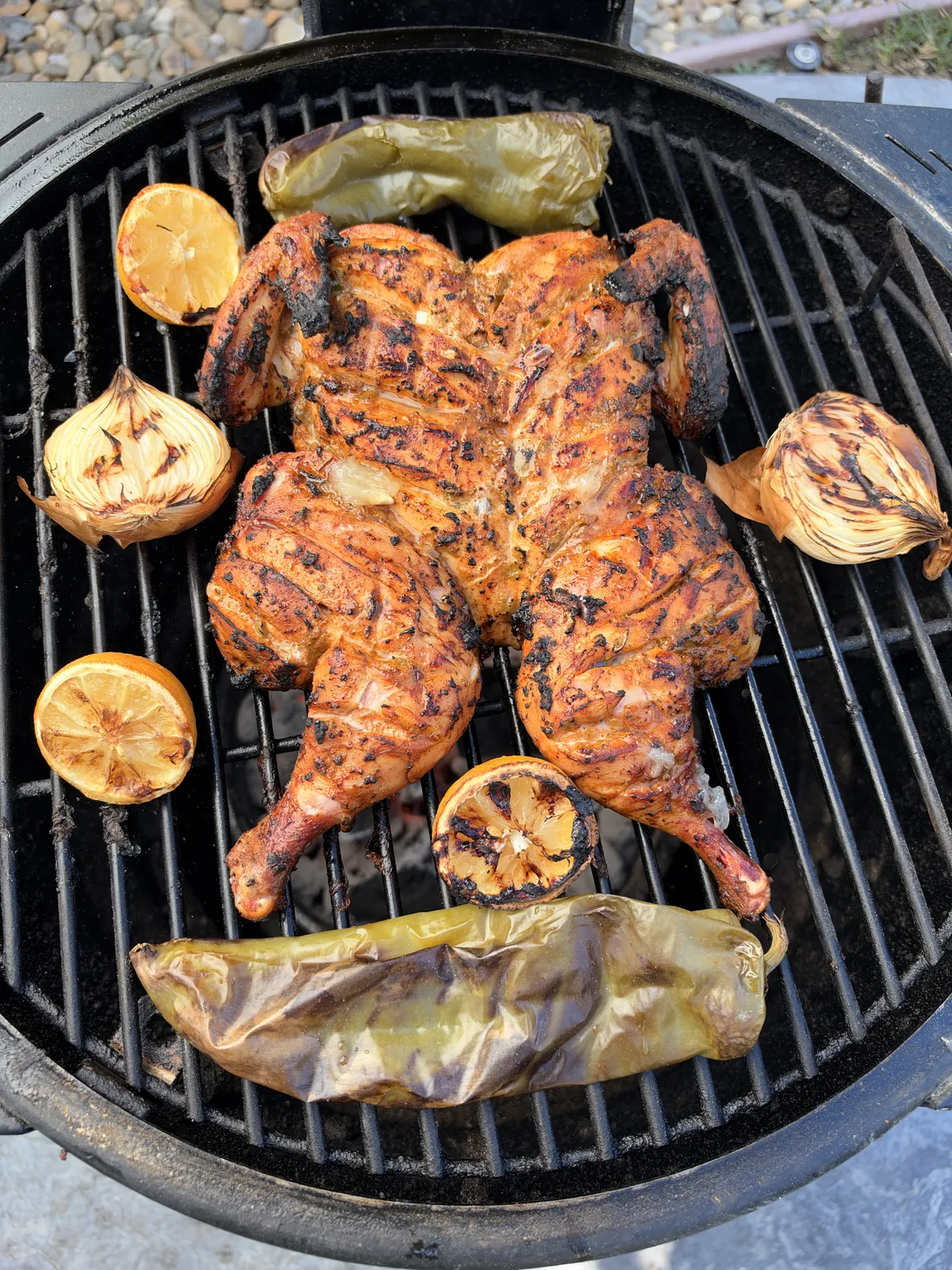 Spatchcocked chicken grilling at 350°F on charcoal grill surrounded by charred onion halves, grilled lemon halves, and roasted Anaheim peppers