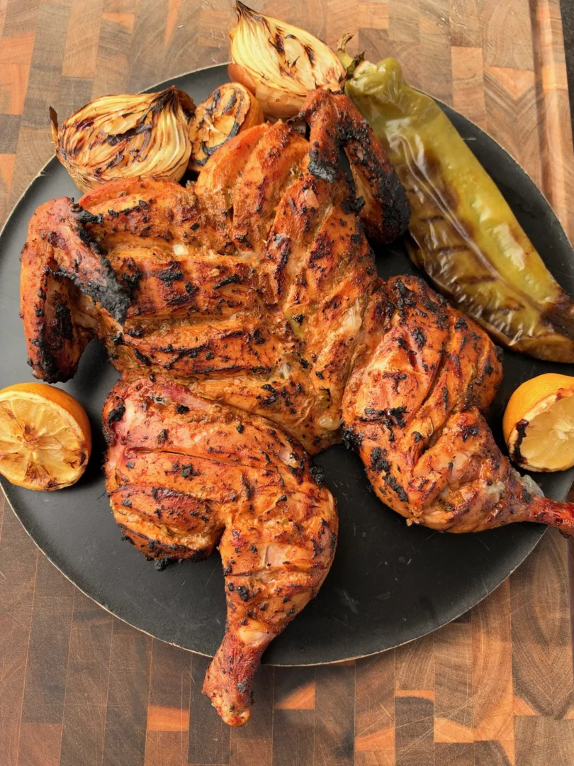Spatchcocked green chile lime grilled chicken with charred lemons, grilled onions, and Anaheim peppers on a dark plate