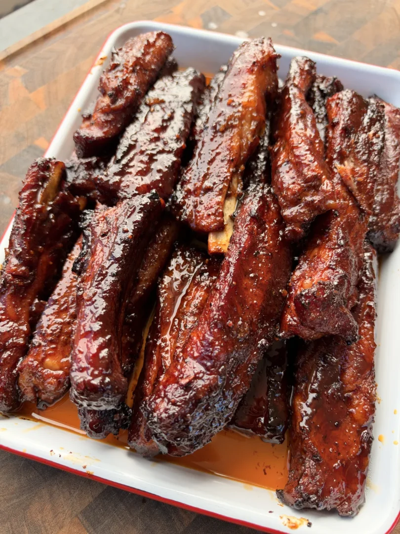 Sticky smoked St. Louis-style barbecue party ribs piled high on a white enamel tray, glazed in a glossy brown sugar honey BBQ sauce