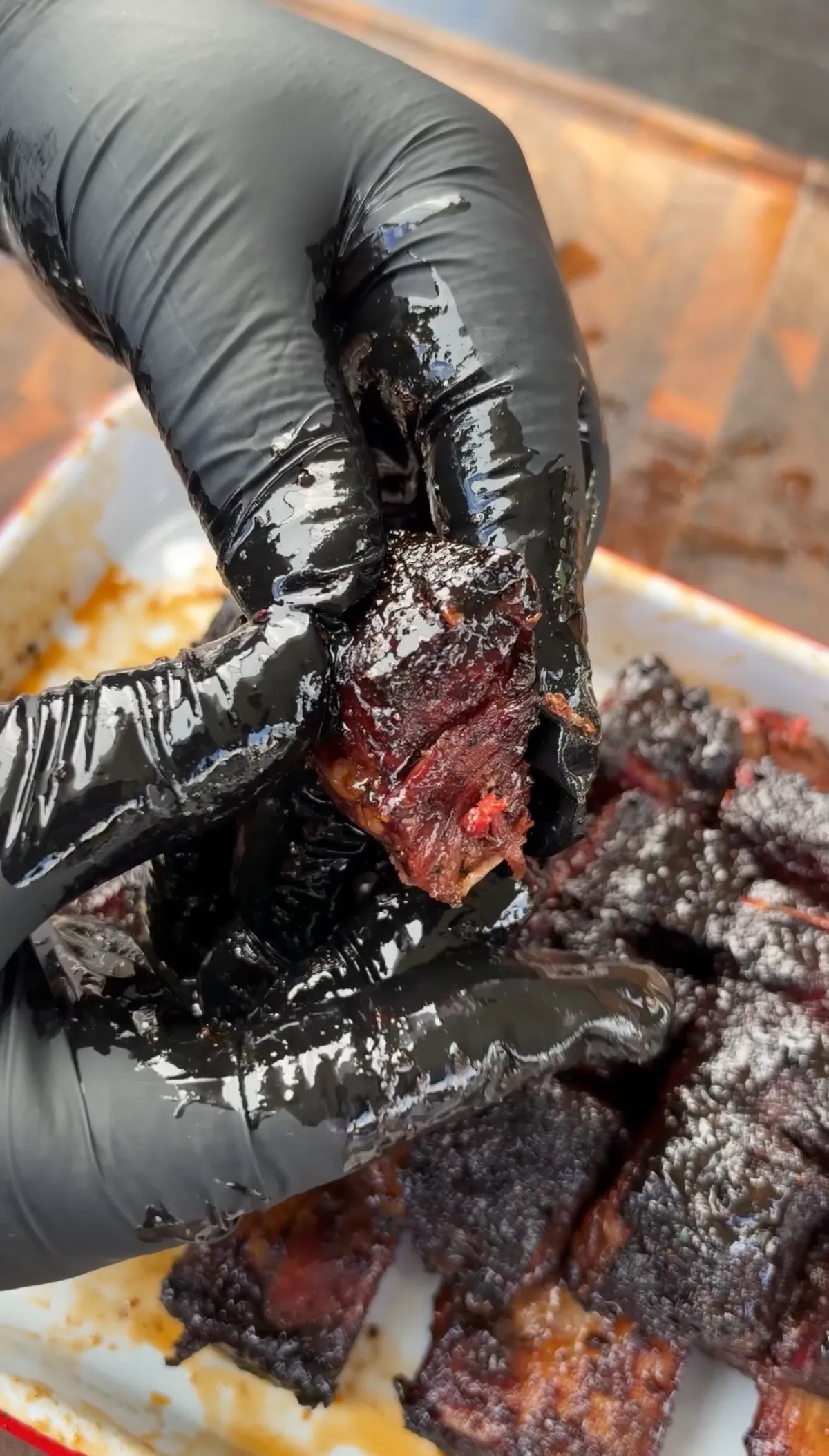 Black-gloved hands pulling apart a sticky, lacquered beef short rib burnt end over a tray of caramelized pieces