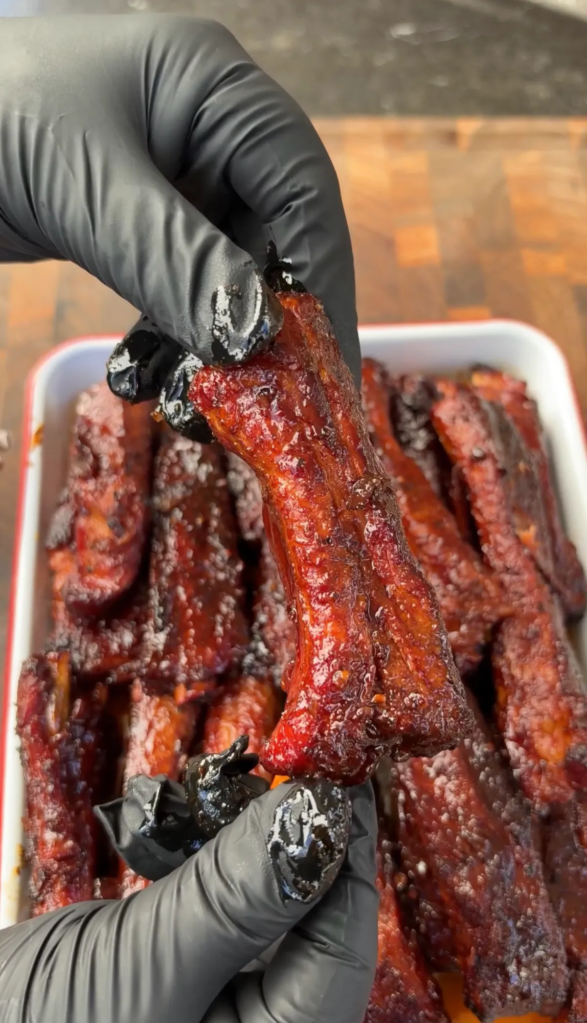Hands in black gloves holding a single glazed St. Louis-style party rib above a tray of sticky smoked barbecue ribs