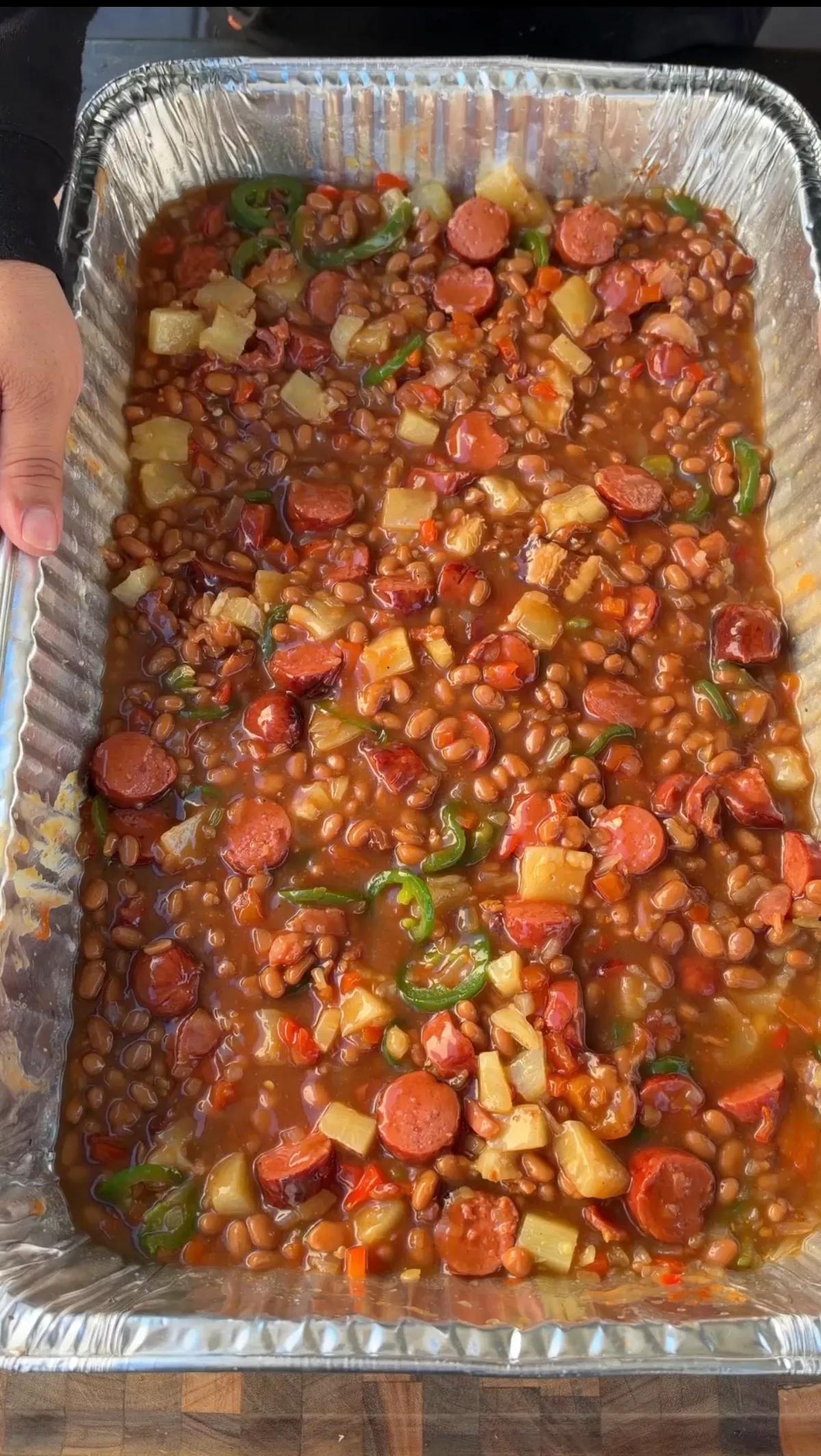 Large foil pan of smoked BBQ baked beans with hot dog rounds, jalapeño slices, pineapple tidbits, and bacon in a rich sauce