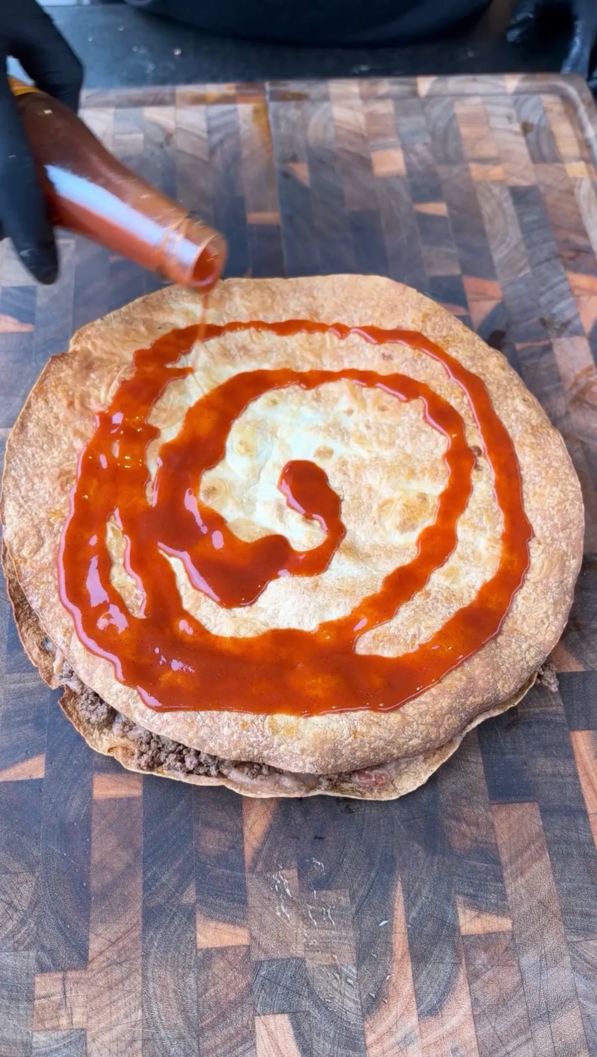 Taco Bell taco sauce being drizzled in a spiral over a crispy flour tortilla layered with seasoned ground beef