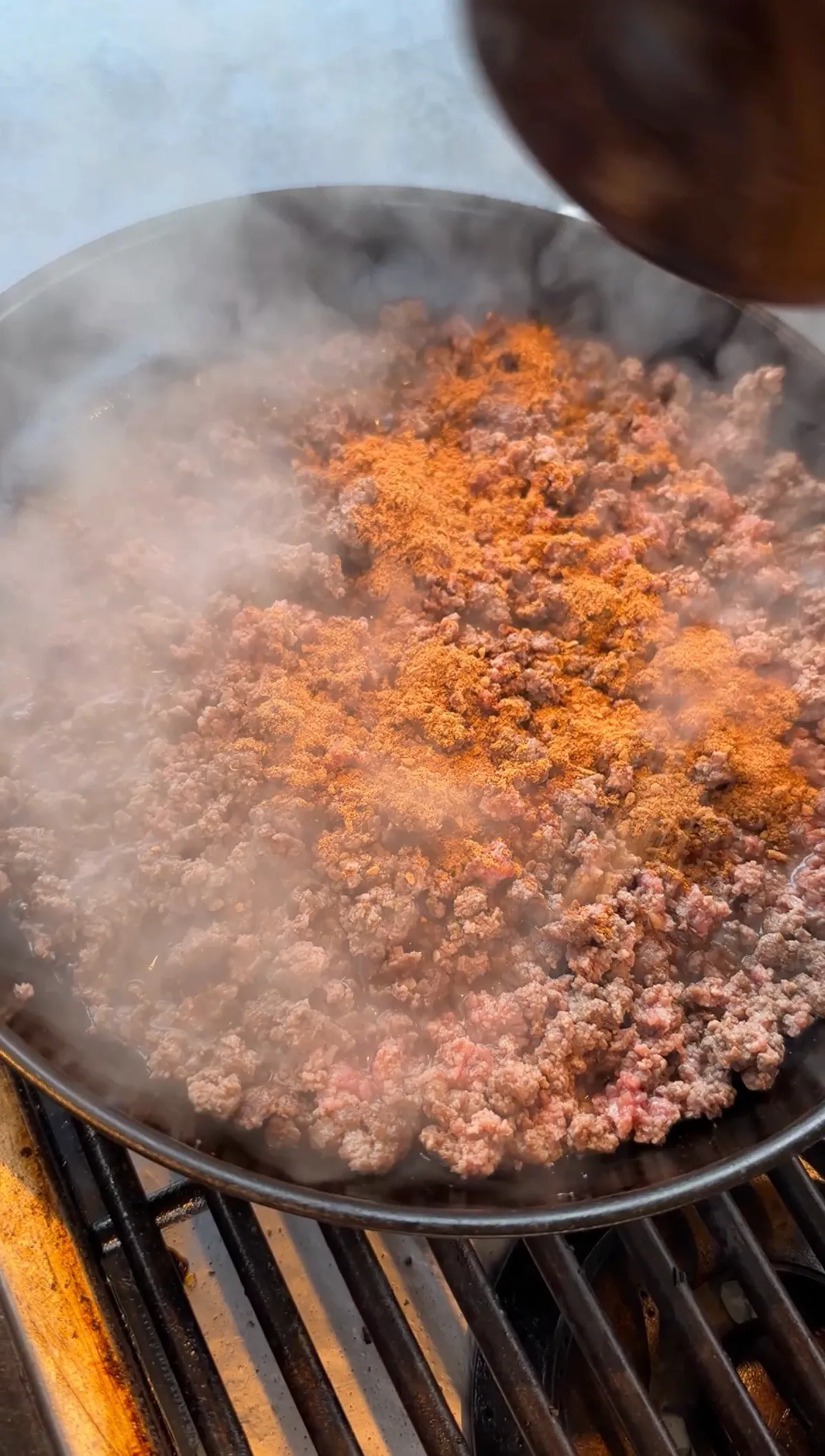Taco seasoning being added to browning 85/15 ground beef in a large skillet on a grill, with steam rising