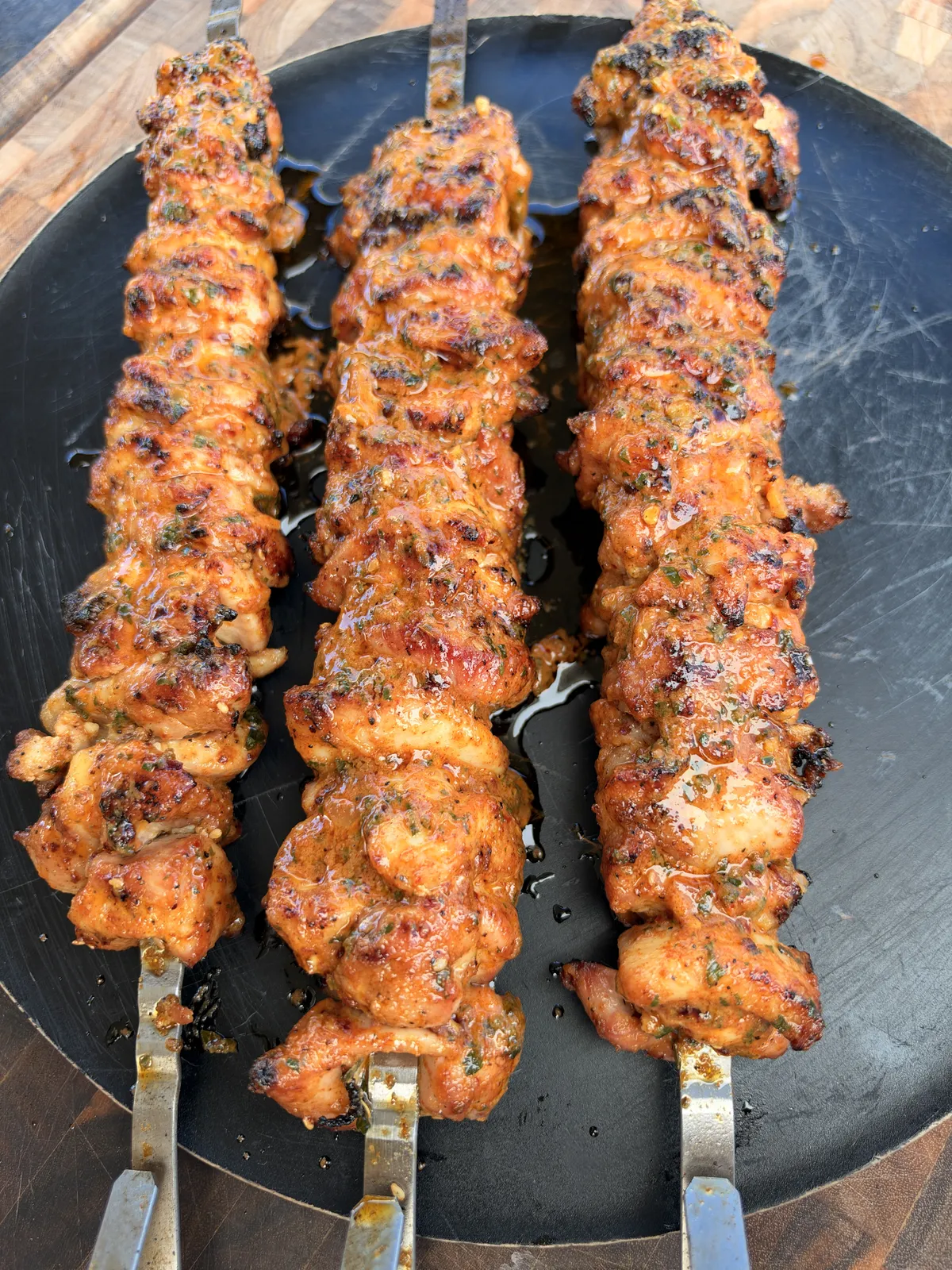 Three grilled chicken thigh skewers basted with cowboy butter resting on a round black metal serving tray