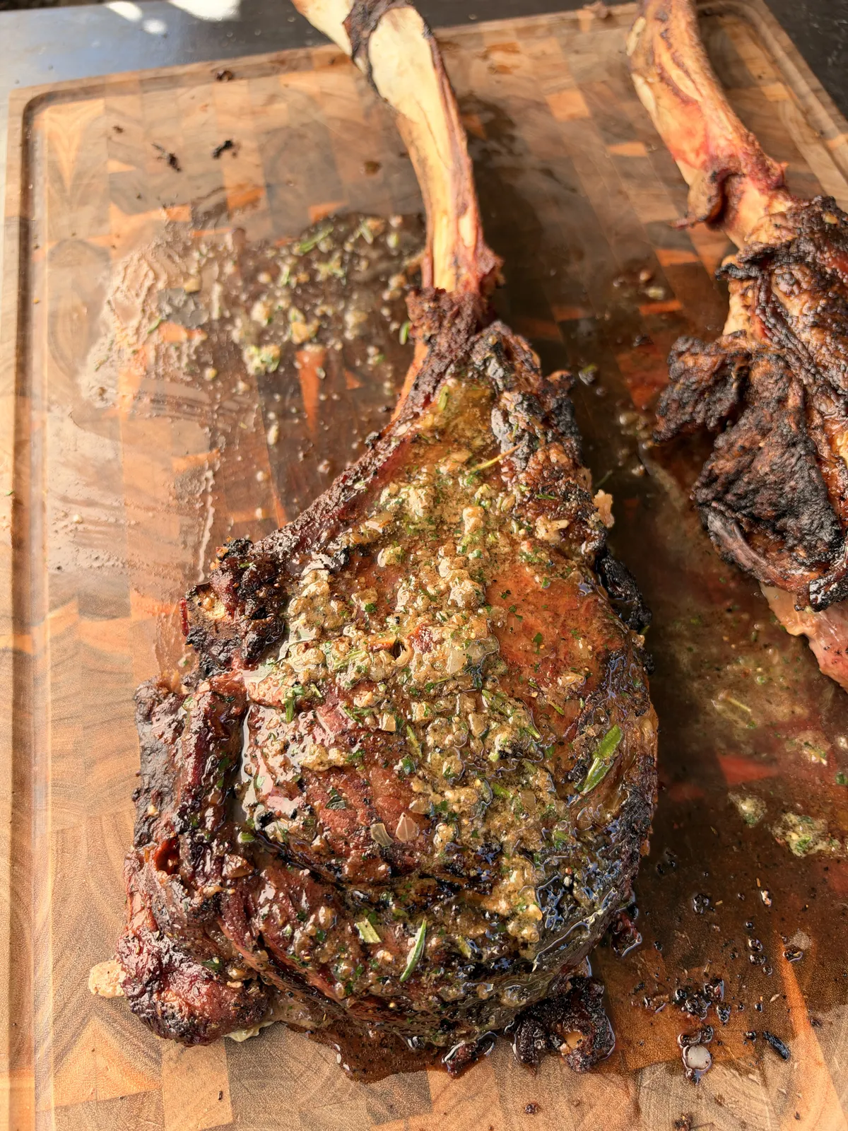 Two-bone prime rib tomahawk topped with roasted garlic and onion compound butter melting over seared crust on wood cutting board