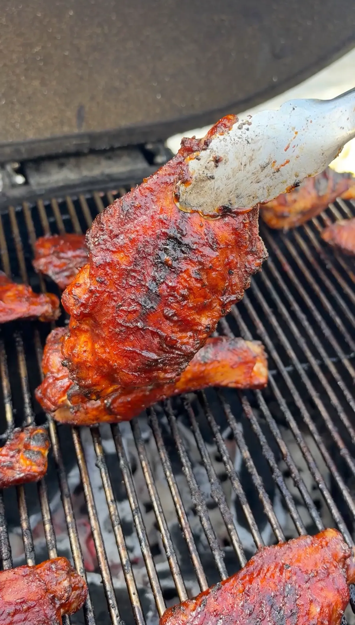 Tongs holding a glazed BBQ turkey wing drumette above charcoal grill grates with caramelized sauce