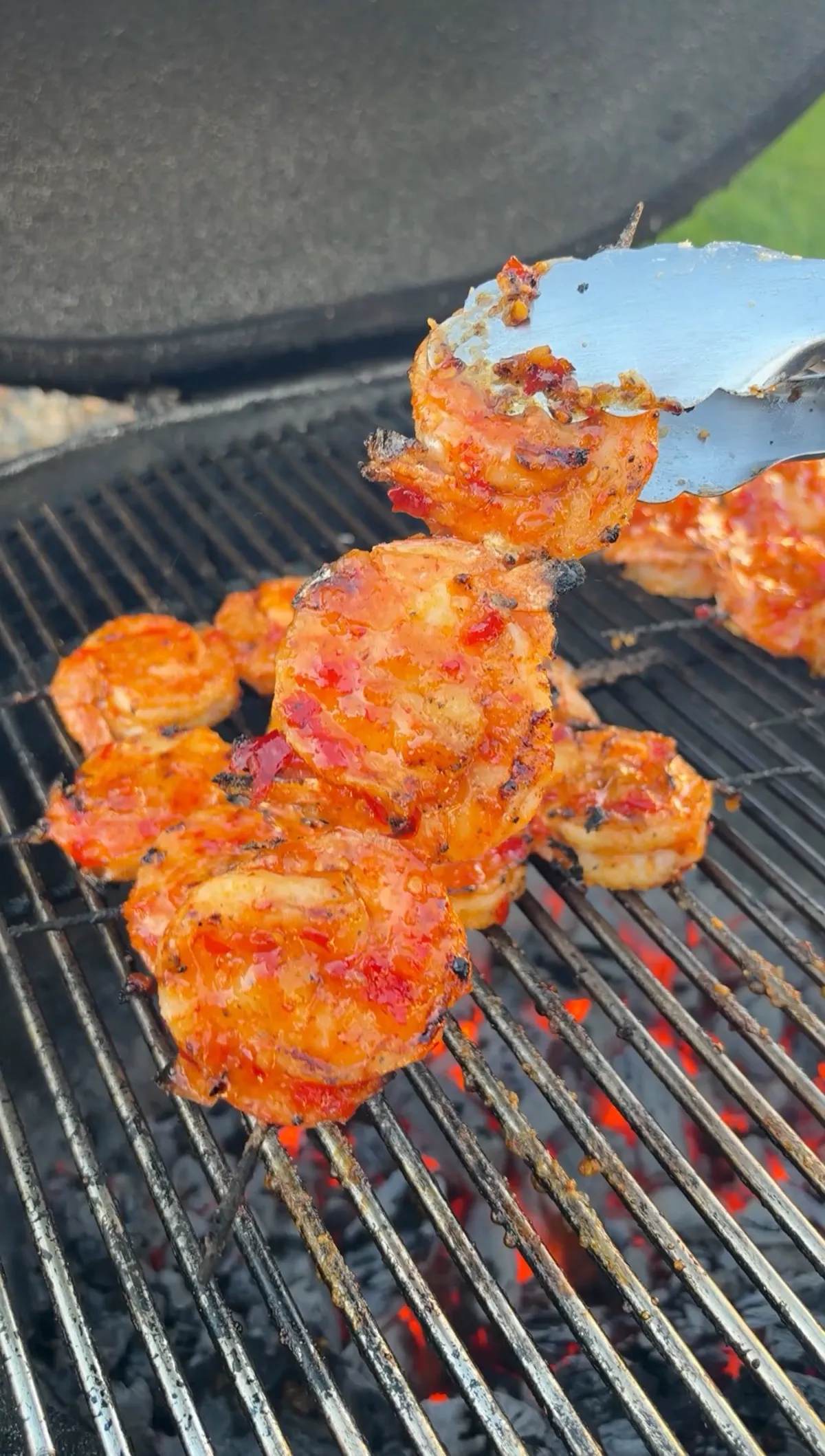 Metal tongs lifting a glazed firecracker shrimp skewer off hot charcoal grill grates with caramelized sweet chili glaze