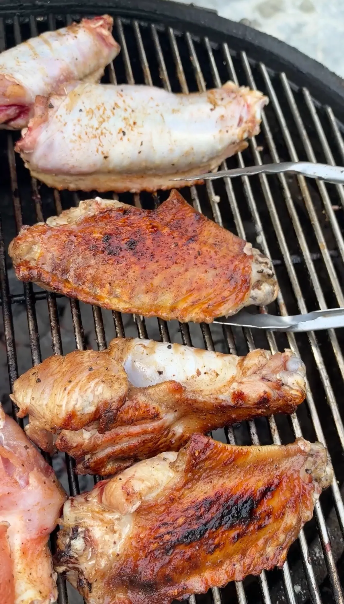 Turkey wing flats and drums at various stages of cooking on charcoal grill, tongs visible, skin browning