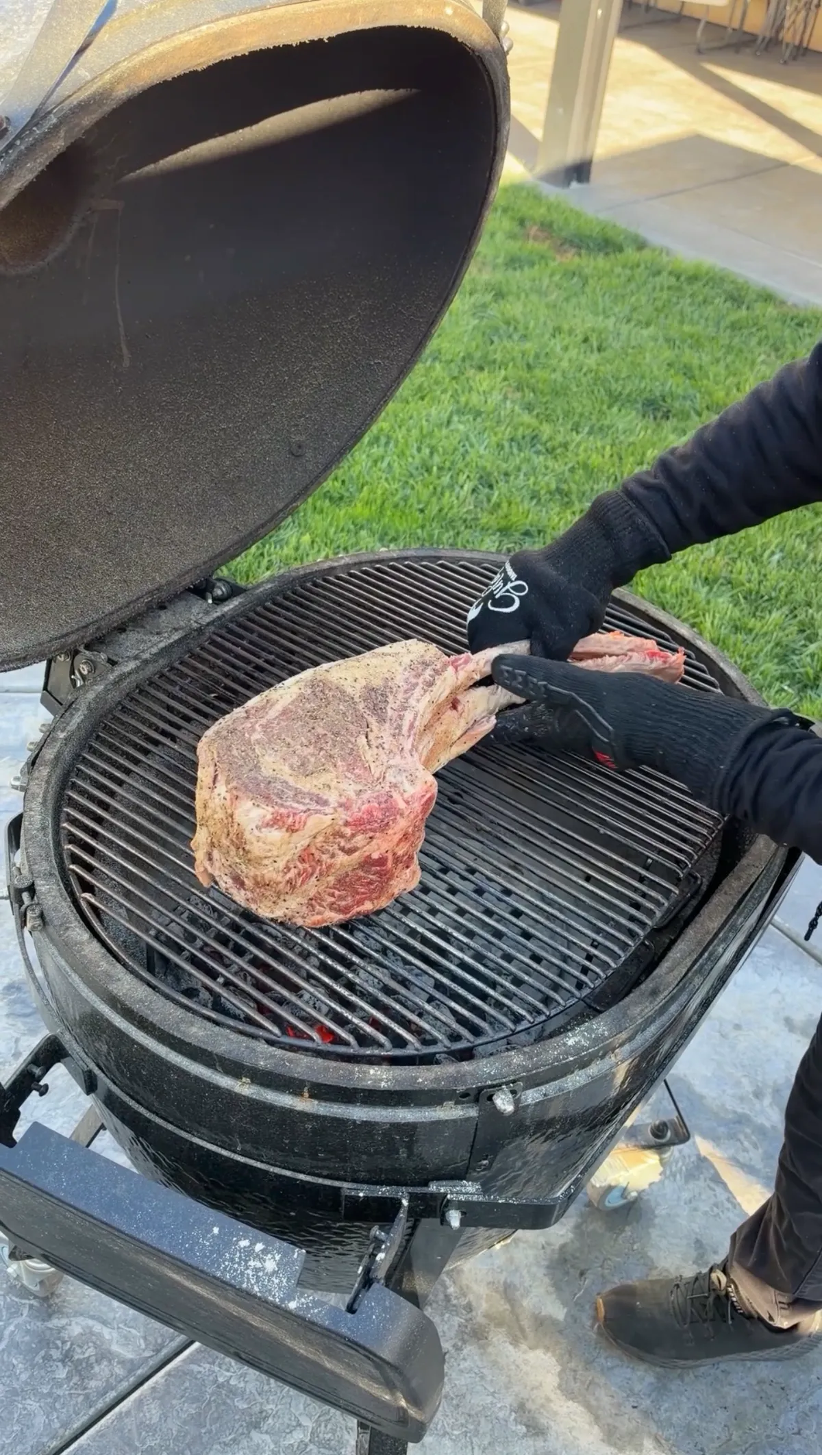 Two-bone prime rib tomahawk being placed on a hot kamado grill grate for forward-searing with gloved hands