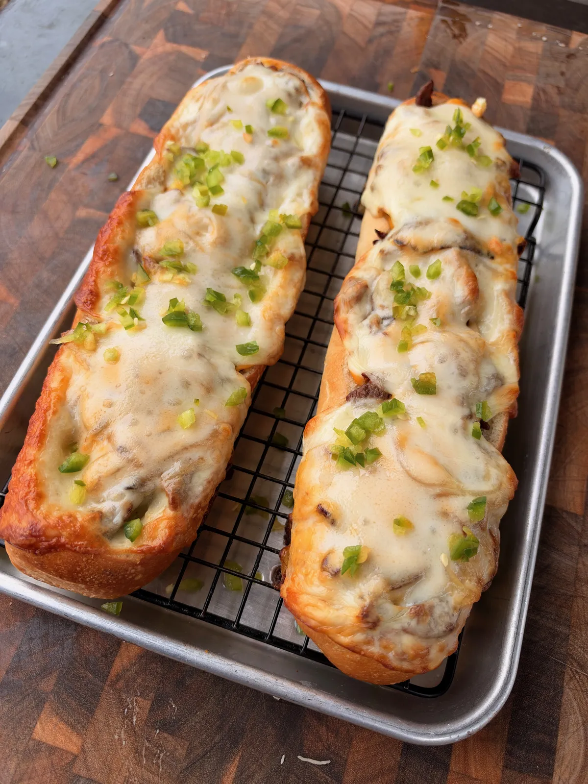 Two baked cheesesteak stuffed baguette halves with melted provolone and diced jalapeños on a wire rack