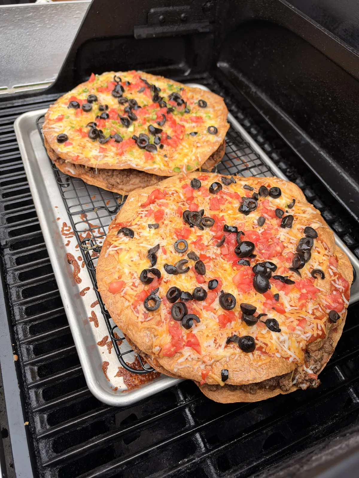 Two Taco Bell copycat Mexican pizzas with melted Colby Jack, diced tomatoes, and sliced black olives on a wire rack baking sheet on a grill